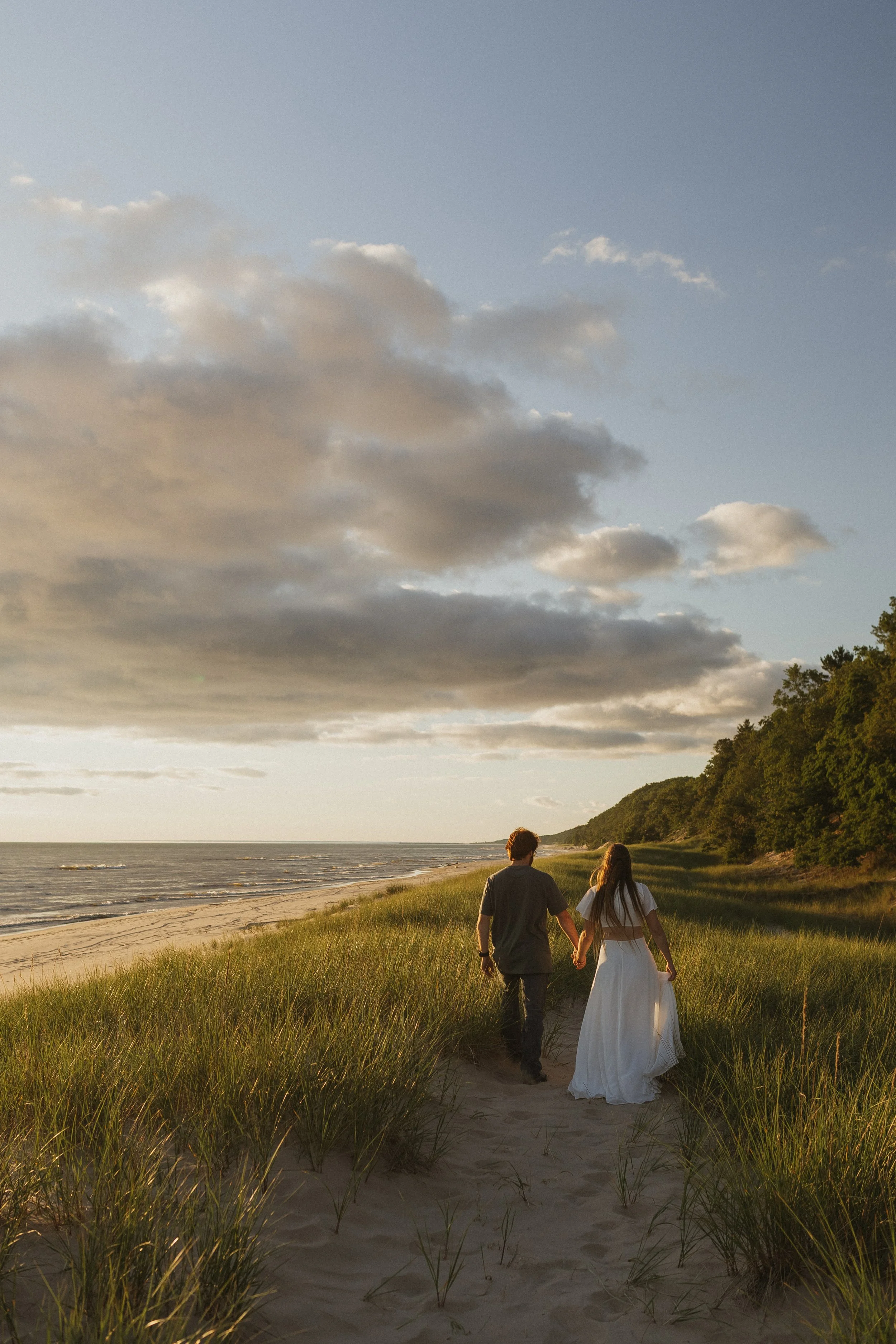 A couple walking hand in hand on a sandy beach at sunset, with grassy dunes and a partly cloudy sky in the background.