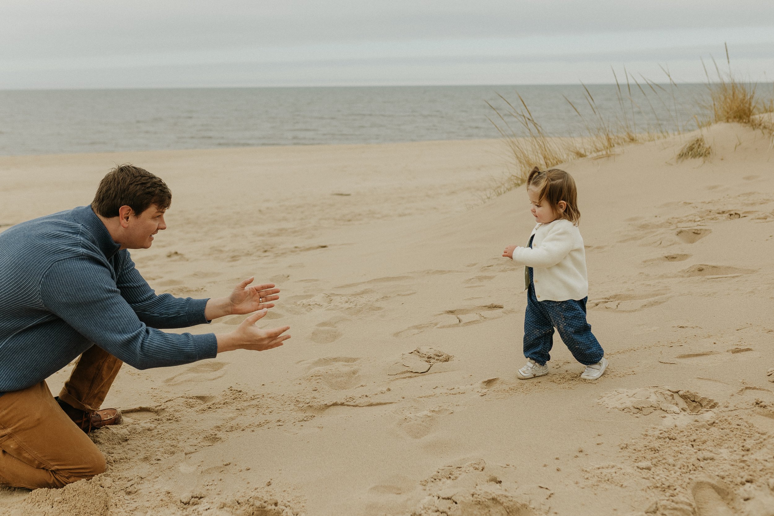 A man kneeling on the sand at the beach, reaching out towards a small girl standing a few feet away, with ocean and dunes in the background.