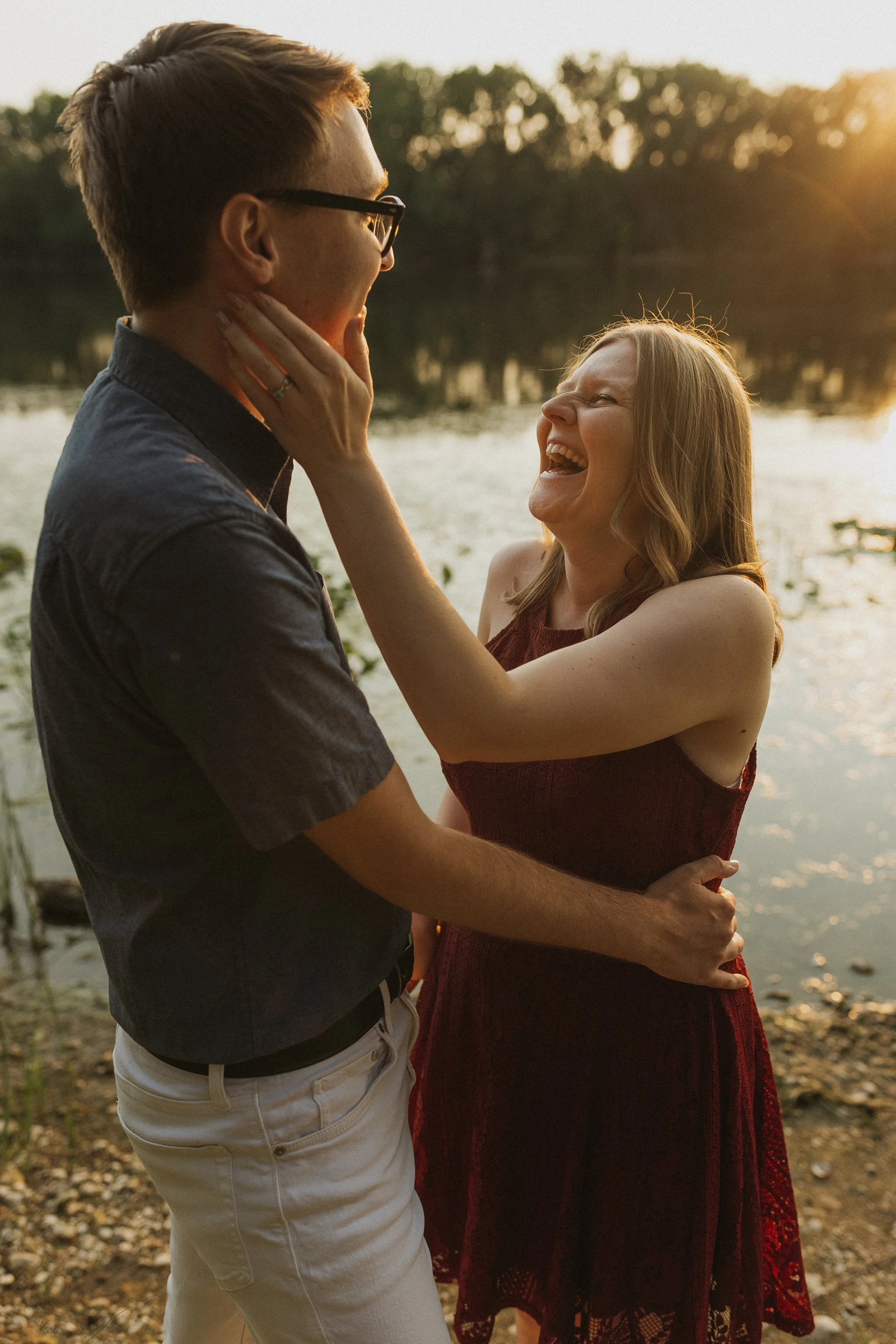 A couple standing near a lakeshore, laughing and holding each other at sunset.