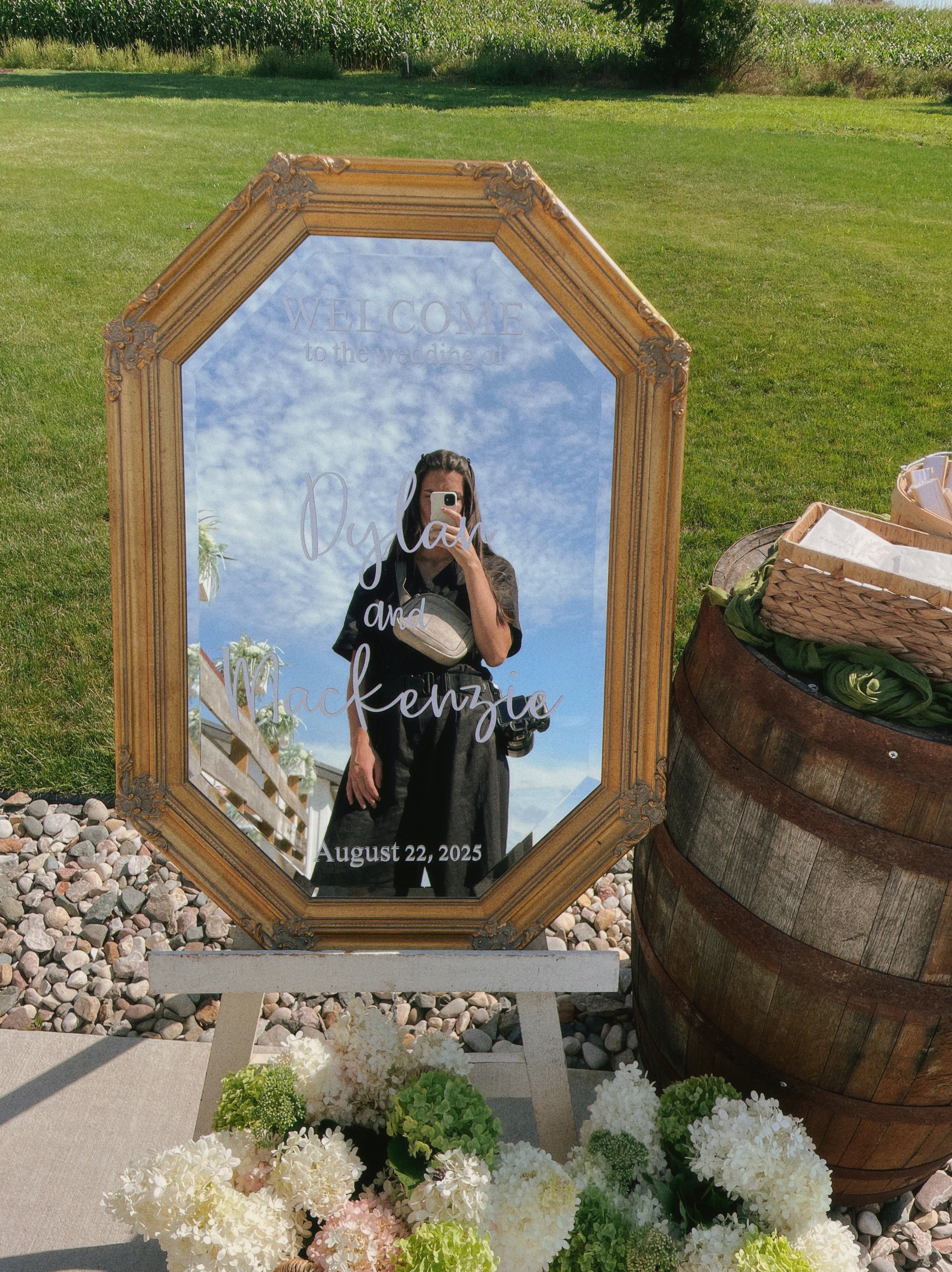 A mirror with a gold ornate frame reflects a woman taking a photo of herself with her phone, with the sky and clouds in the background. The mirror has inscriptions welcoming wedding guests and displaying names 'Oylah and Mackenzie' with the date Augu
