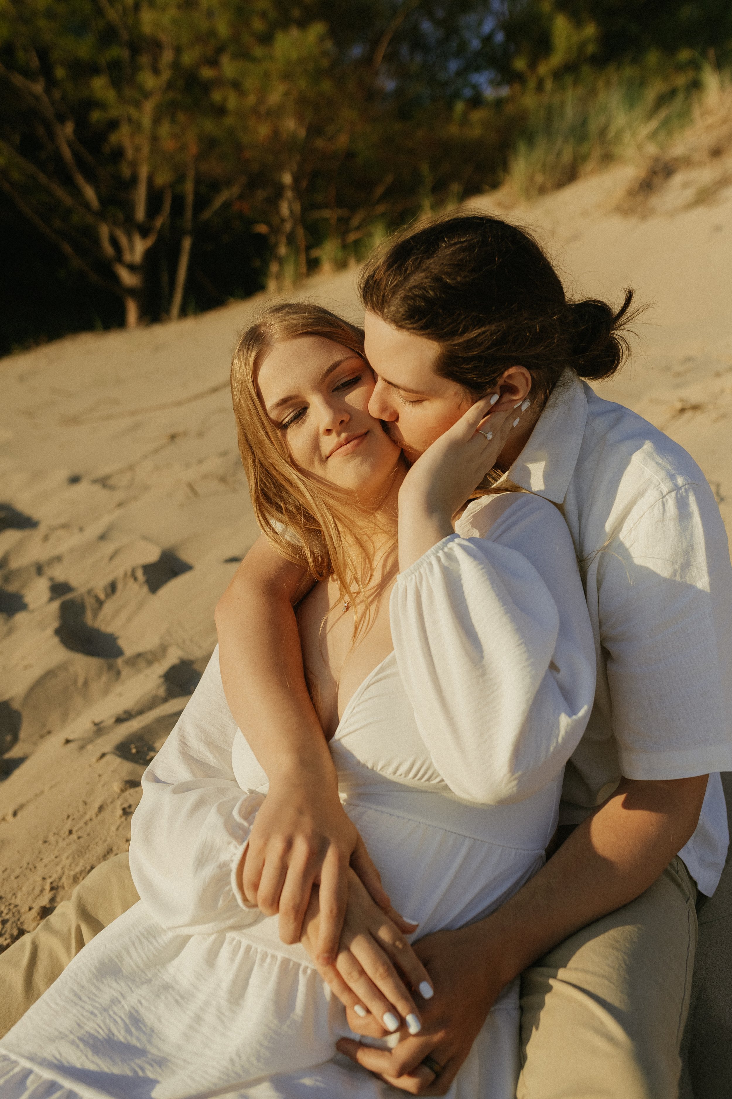 A couple sitting on the sand at the beach, embracing and sharing a tender moment, with trees in the background.