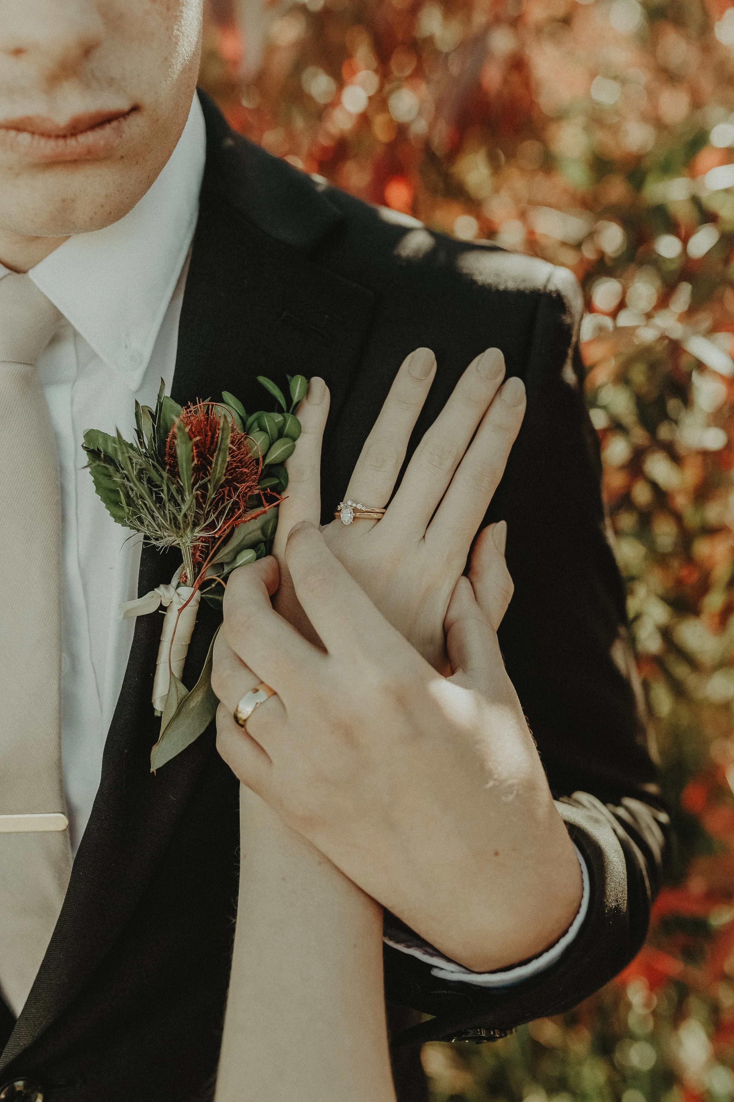 Close-up of a couple's hands showing wedding rings, with the bride's hand resting on the groom's chest. The man is wearing a dark suit with a boutonniere, and the woman’s hand displays an engagement ring and wedding band.