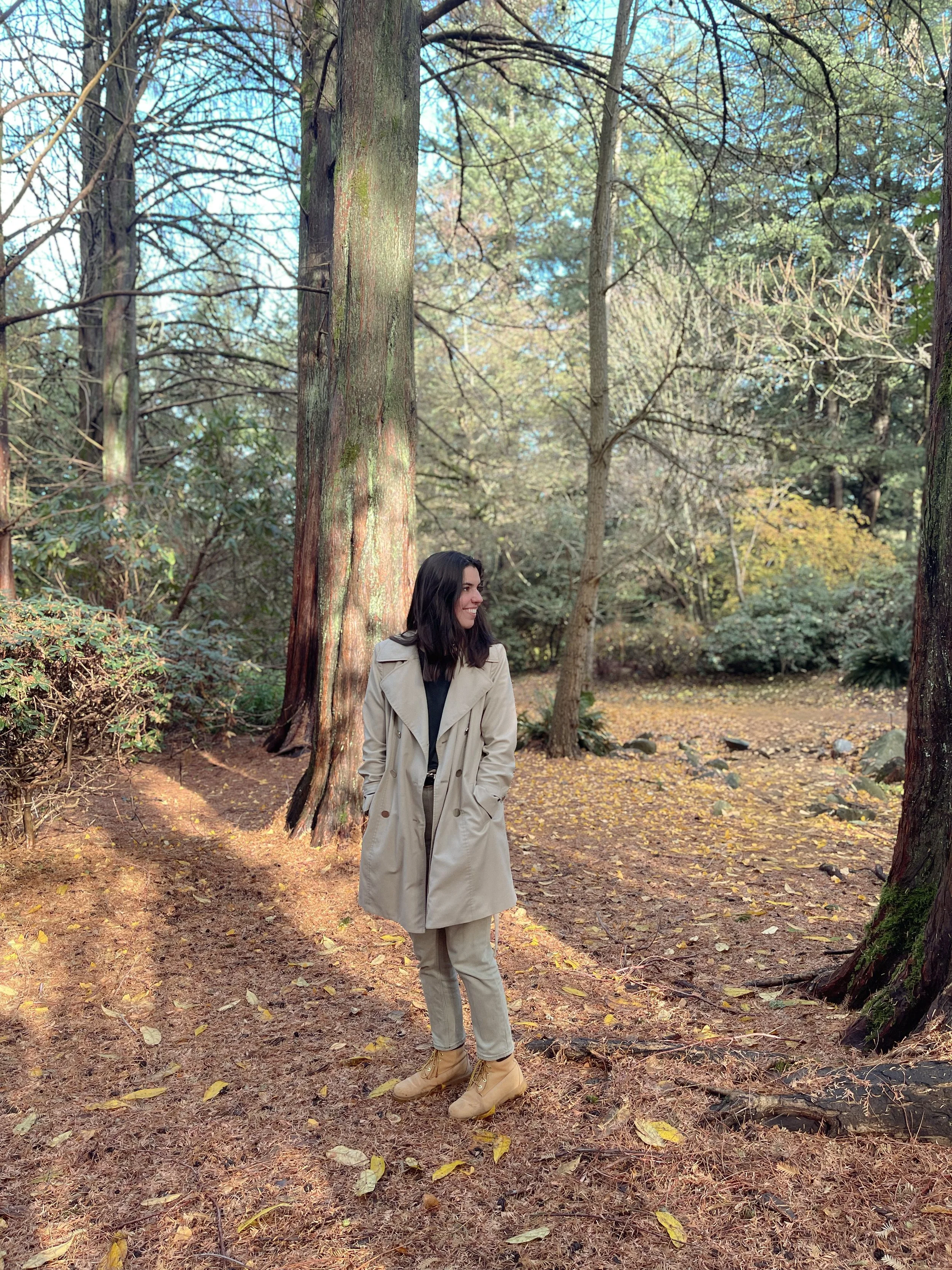 A woman smiling and walking in a forest with tall trees and fallen leaves on the ground, wearing a beige trench coat, beige boots, and beige pants.
