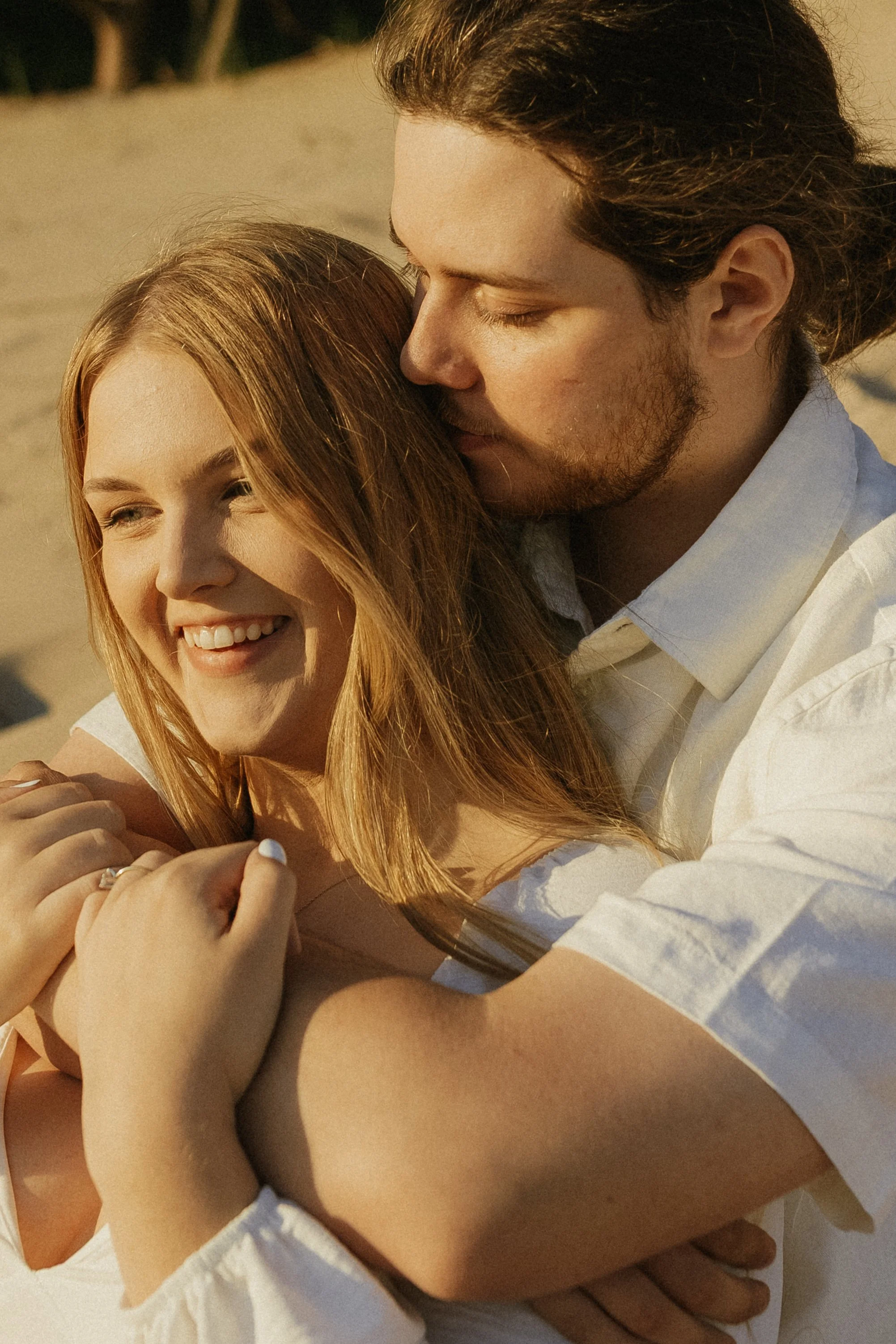 A couple embracing on the beach, the man is kissing the woman's head while she smiles and looks away.