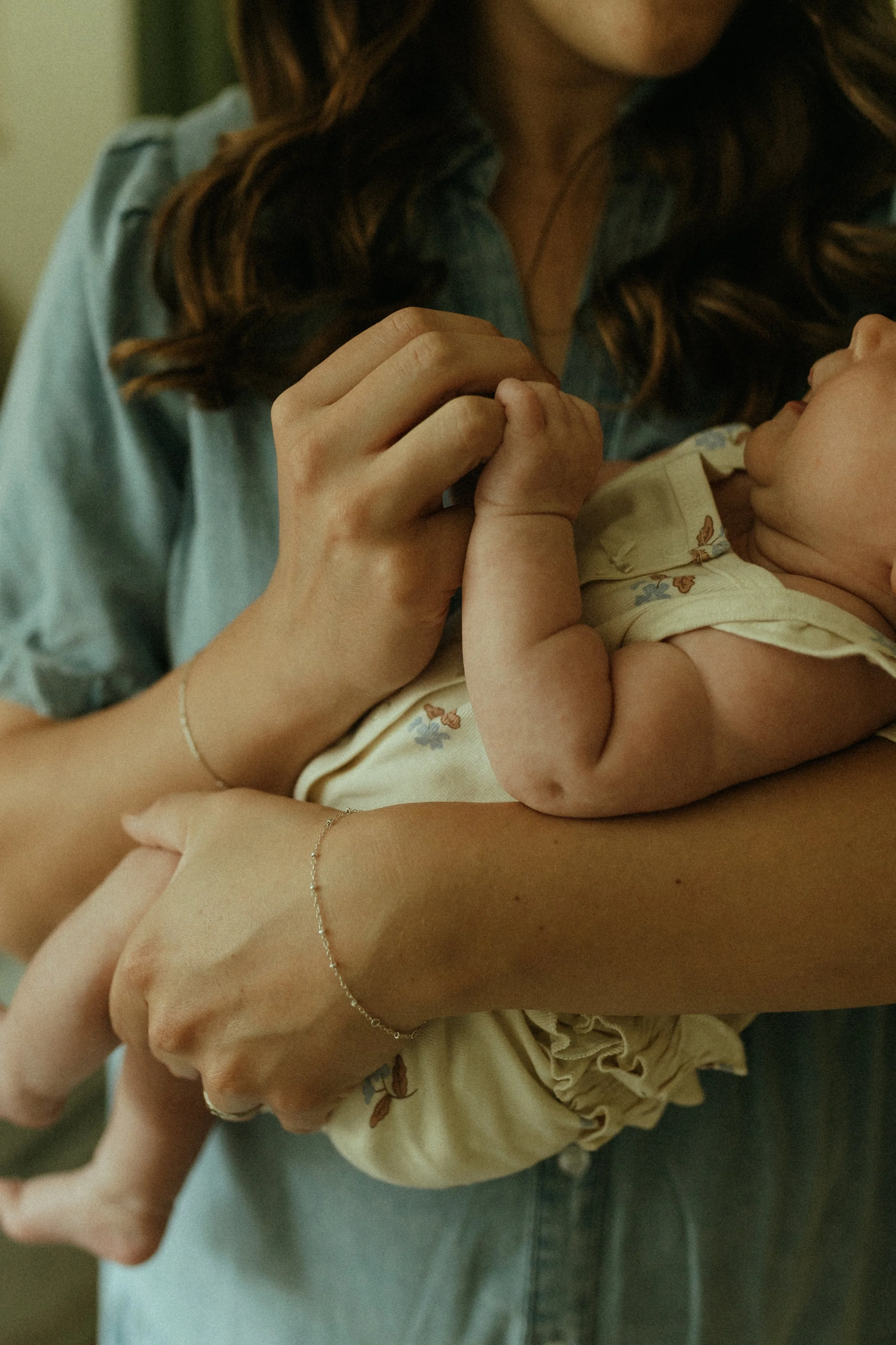 A woman holding a baby close, gently touching the baby's hand. The woman is wearing a blue denim shirt and silver jewelry. The baby is dressed in cream-colored clothing with small floral patterns.