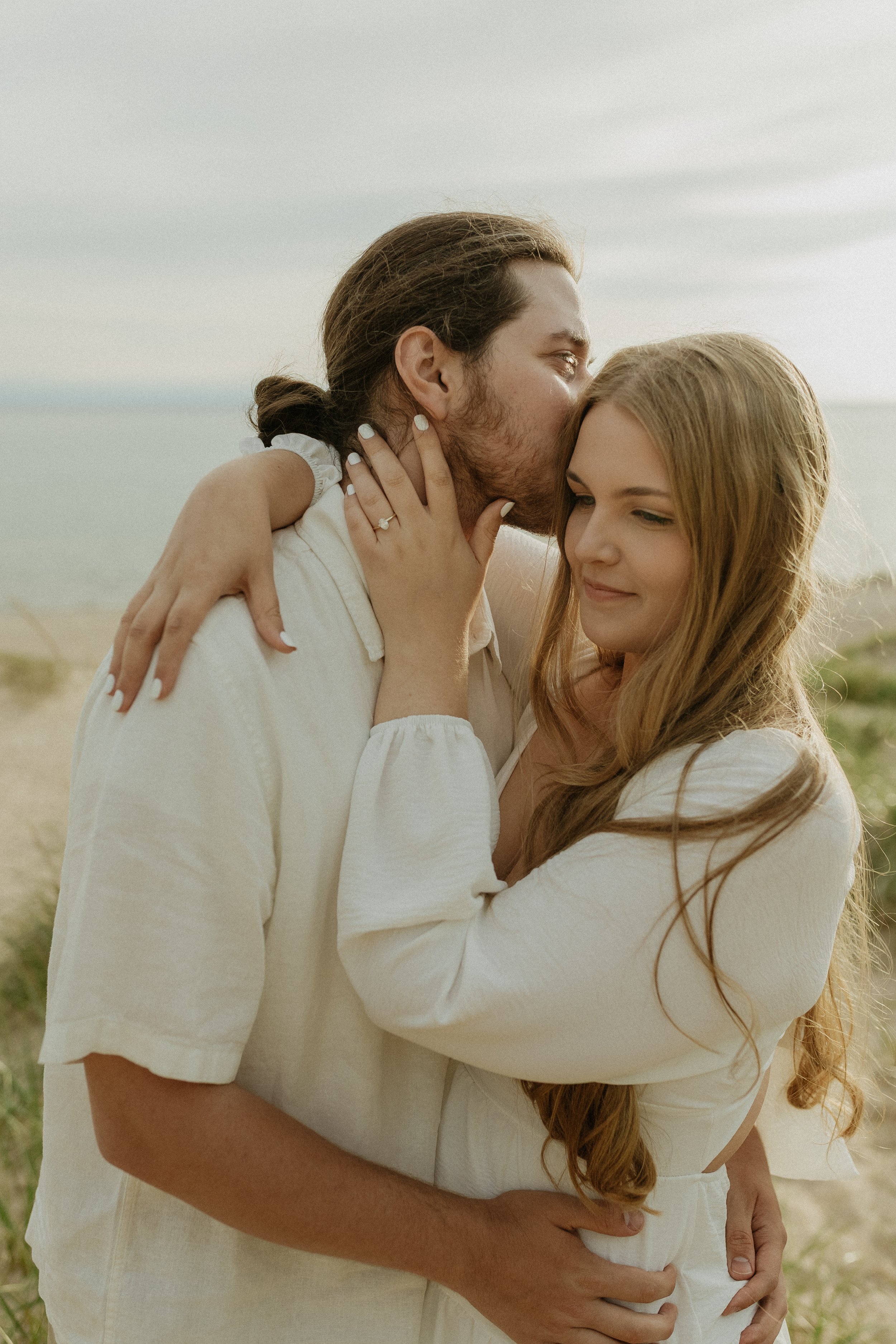 A couple embraces on a beach, with the man kissing the woman's forehead and the woman smiling softly.