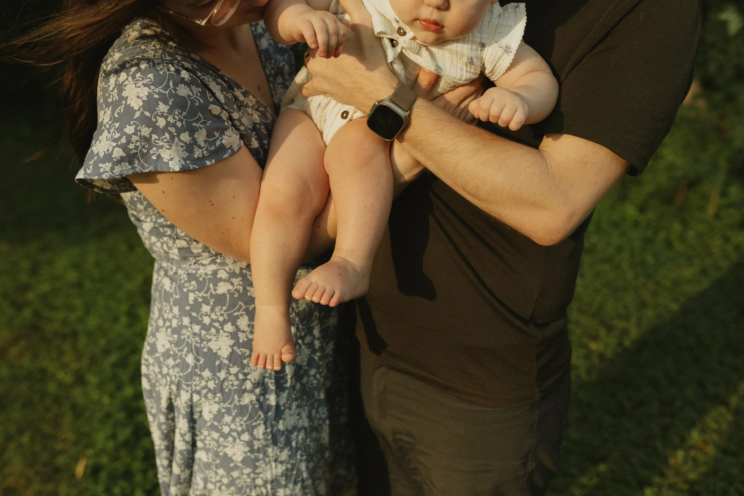 Close-up of a family holding a young child outdoors on grass during the evening, focusing on the baby's feet.