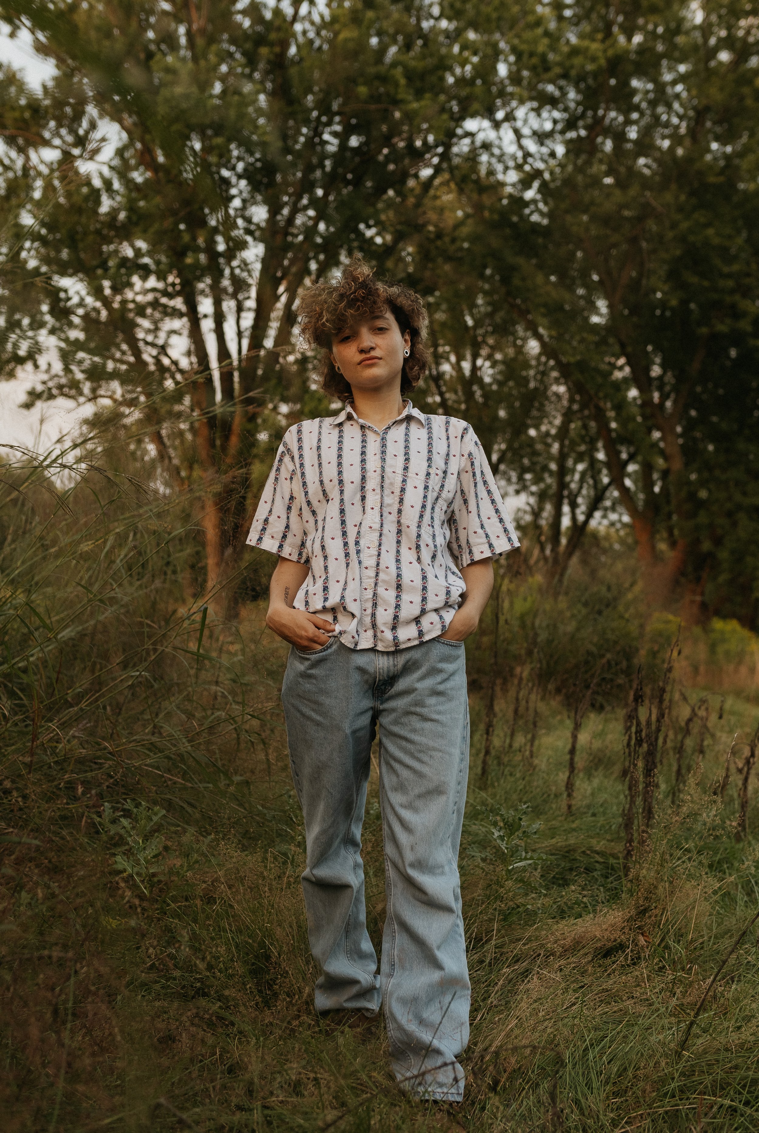Young person wearing a striped shirt and jeans, standing in a grassy field with trees in the background.
