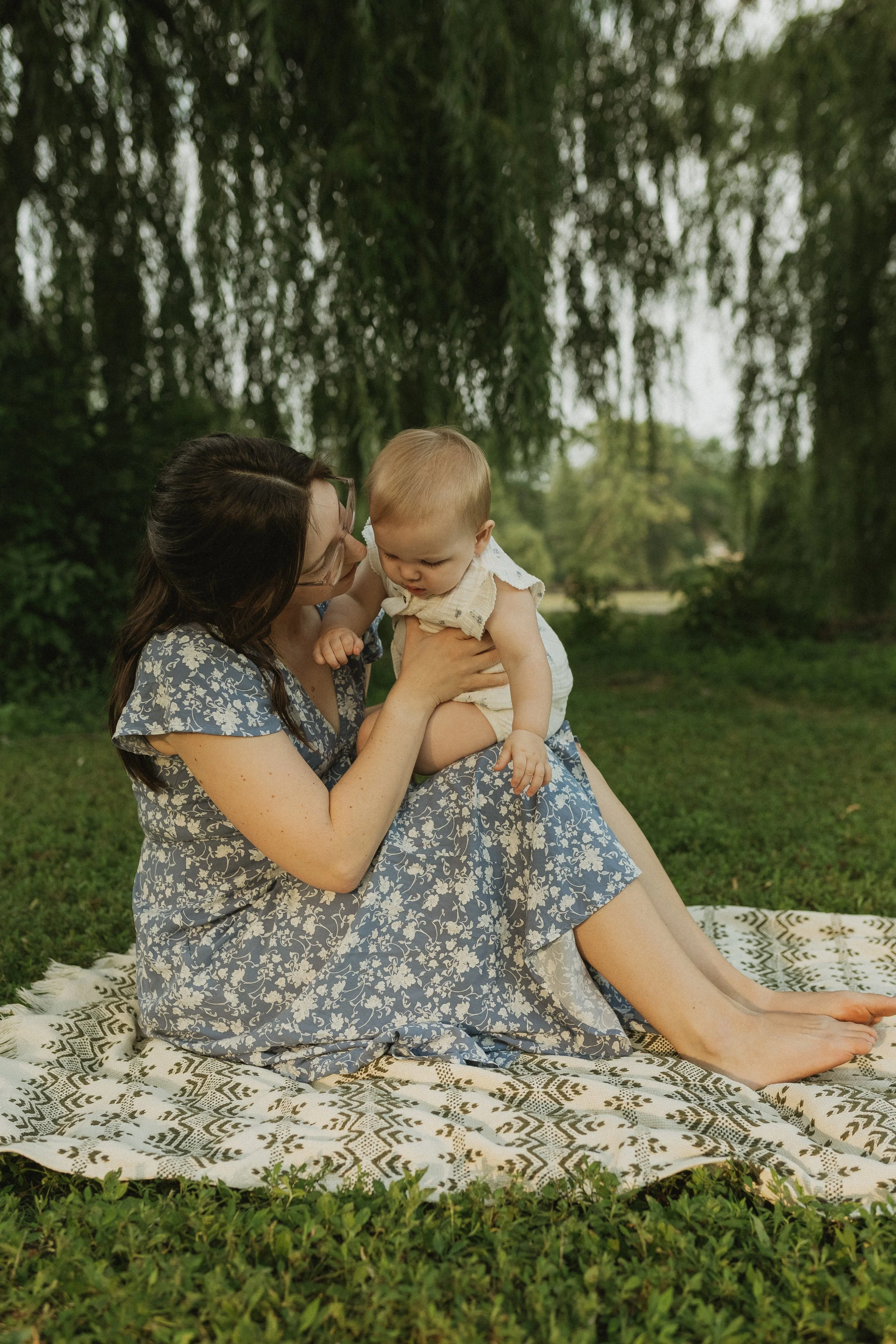 A woman with long dark hair, wearing glasses and a floral dress, sitting on a patterned blanket outdoors, holding a small toddler boy dressed in a light-colored outfit, with trees and greenery in the background.