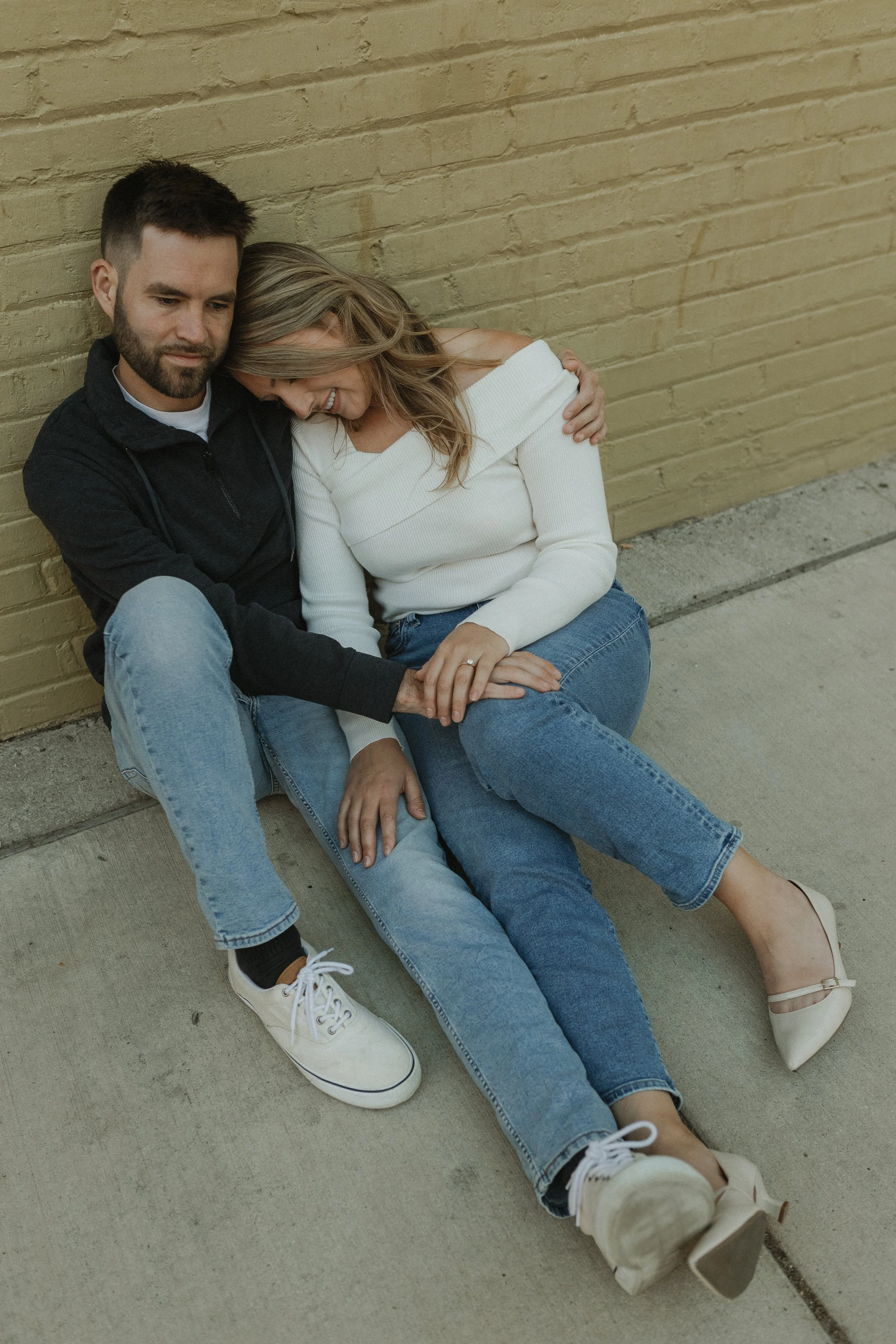 A man and woman sitting on the sidewalk leaning against a yellow brick wall, holding hands and smiling