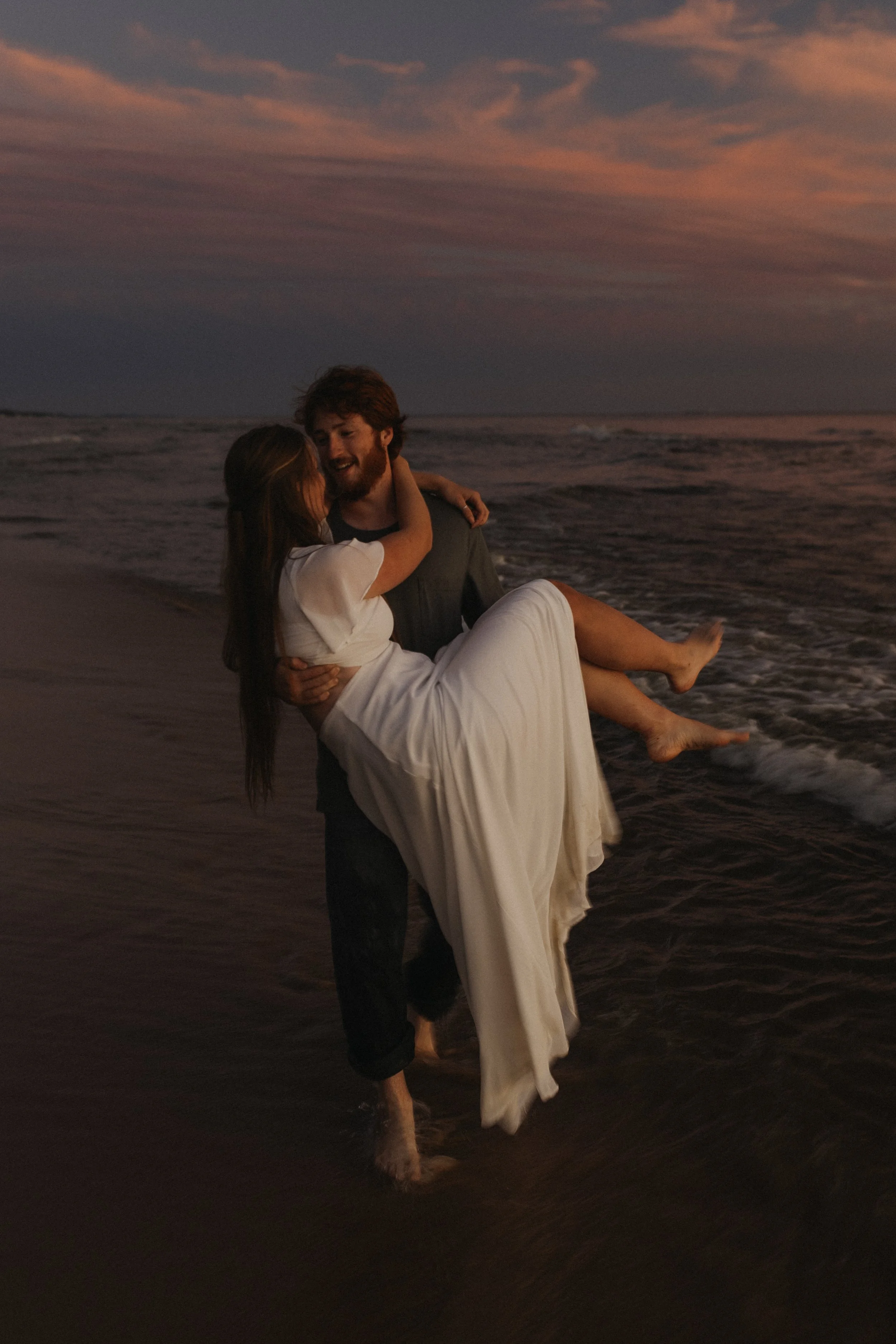 A man holding a woman in a white dress on a beach during sunset.