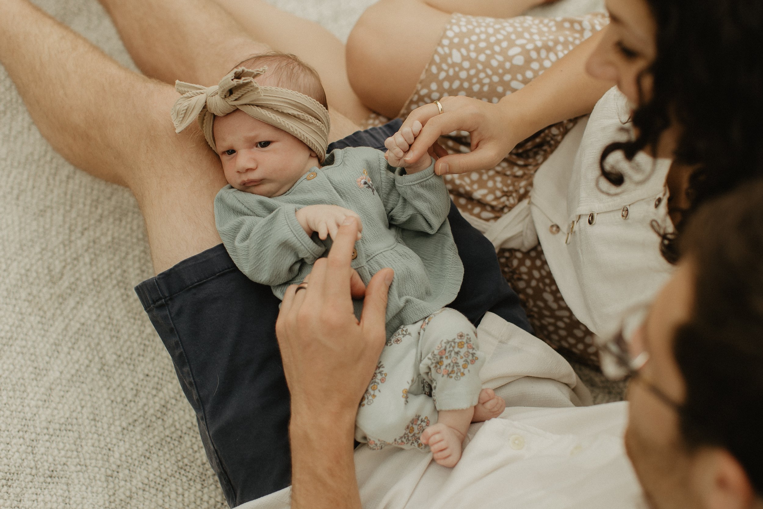 Newborn baby lying on father's lap and holding his finger, wearing a headband and green outfit, while the mother sits next to the father and holds the baby's other hand.