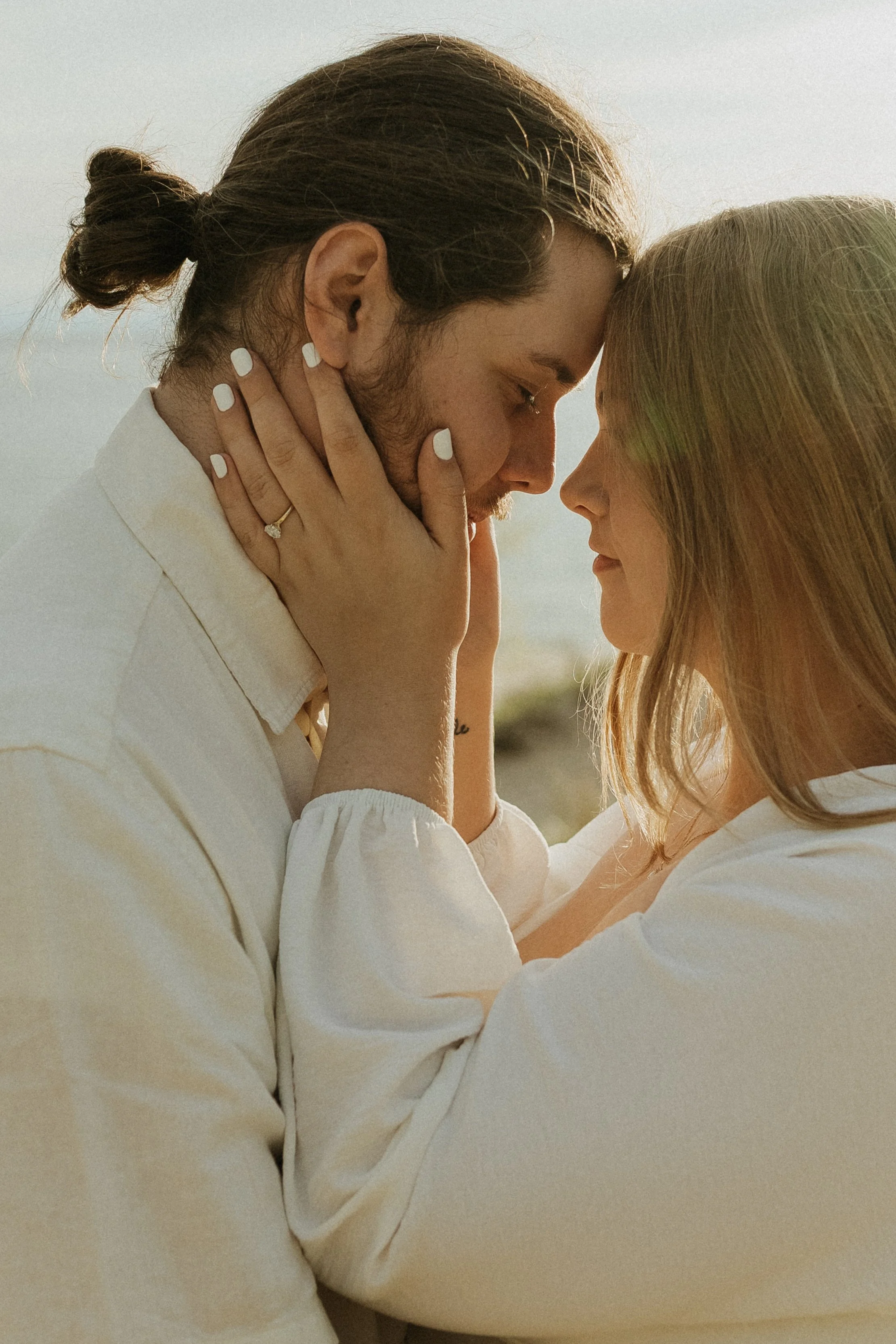 A close-up of a couple wearing all white, with their foreheads touching, holding each other's faces, with a soft, natural background.