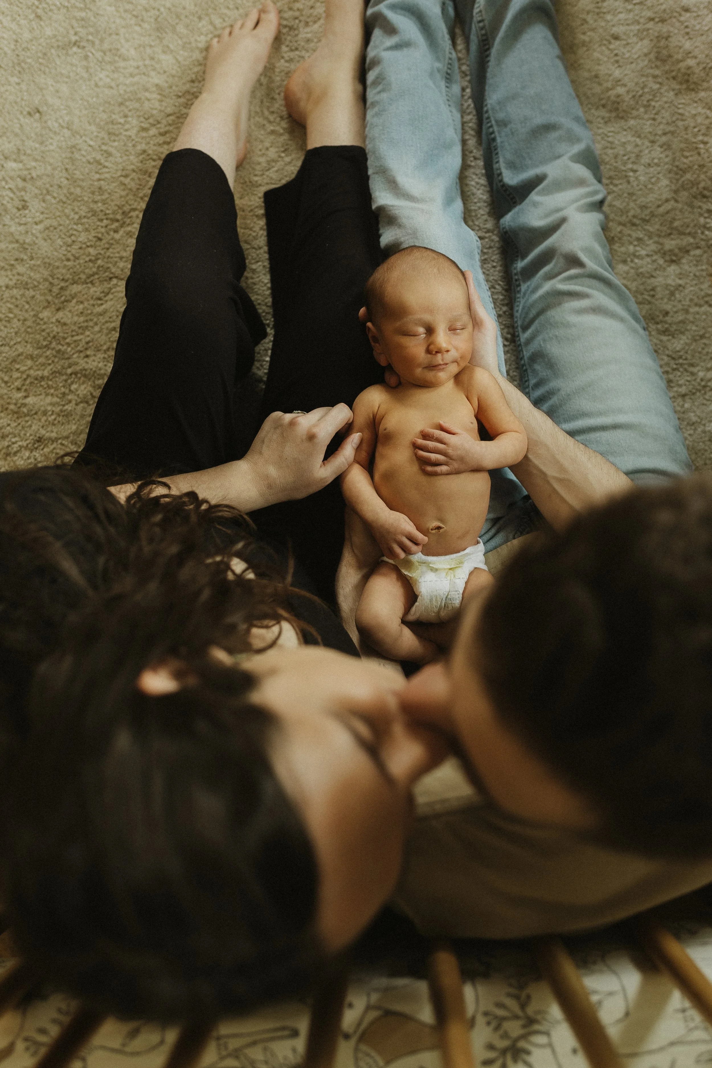 A family of four sitting on the floor, with a newborn baby in the center, adults kissing and touching the baby, on a beige carpet.
