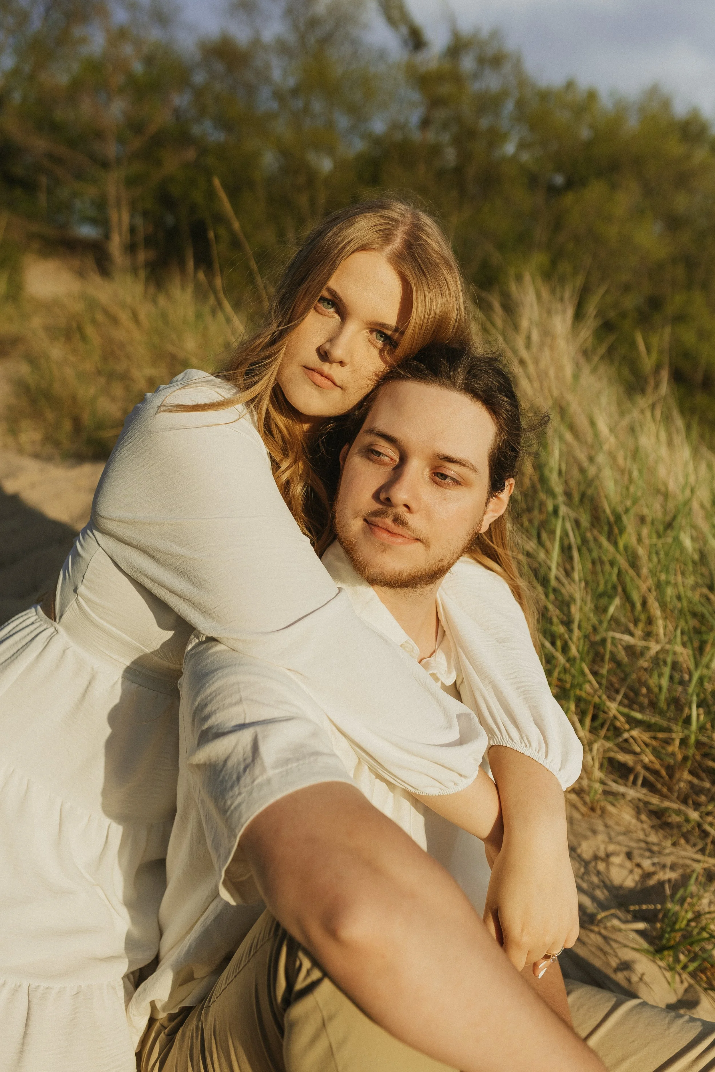A young couple sitting in a grassy outdoor area during golden hour, embracing each other and looking off into the distance.