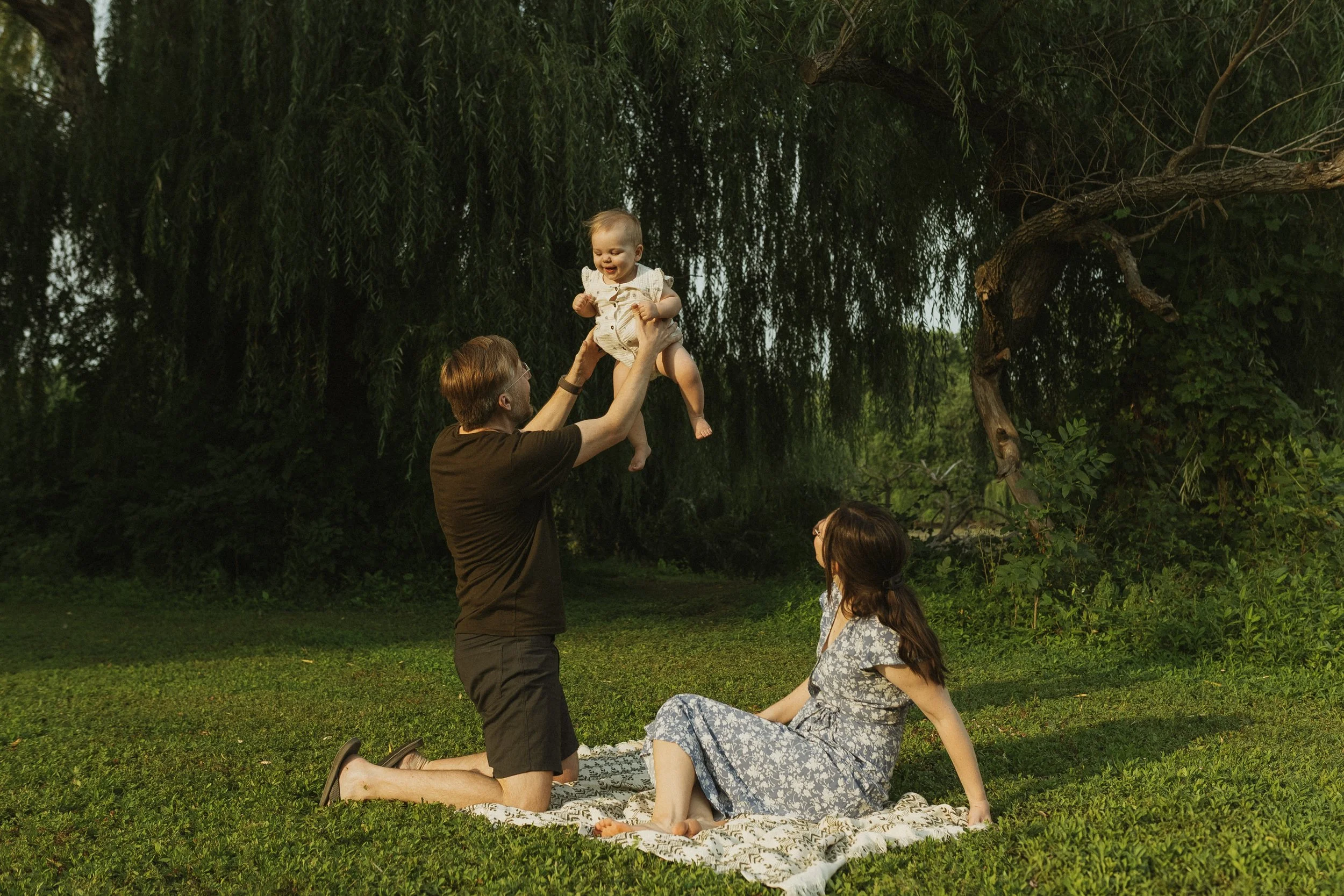 A family enjoying a picnic outdoors. A man holding a smiling baby girl above his head while a woman sits on a blanket on the grass, looking up at them. There is a large leafy tree in the background.