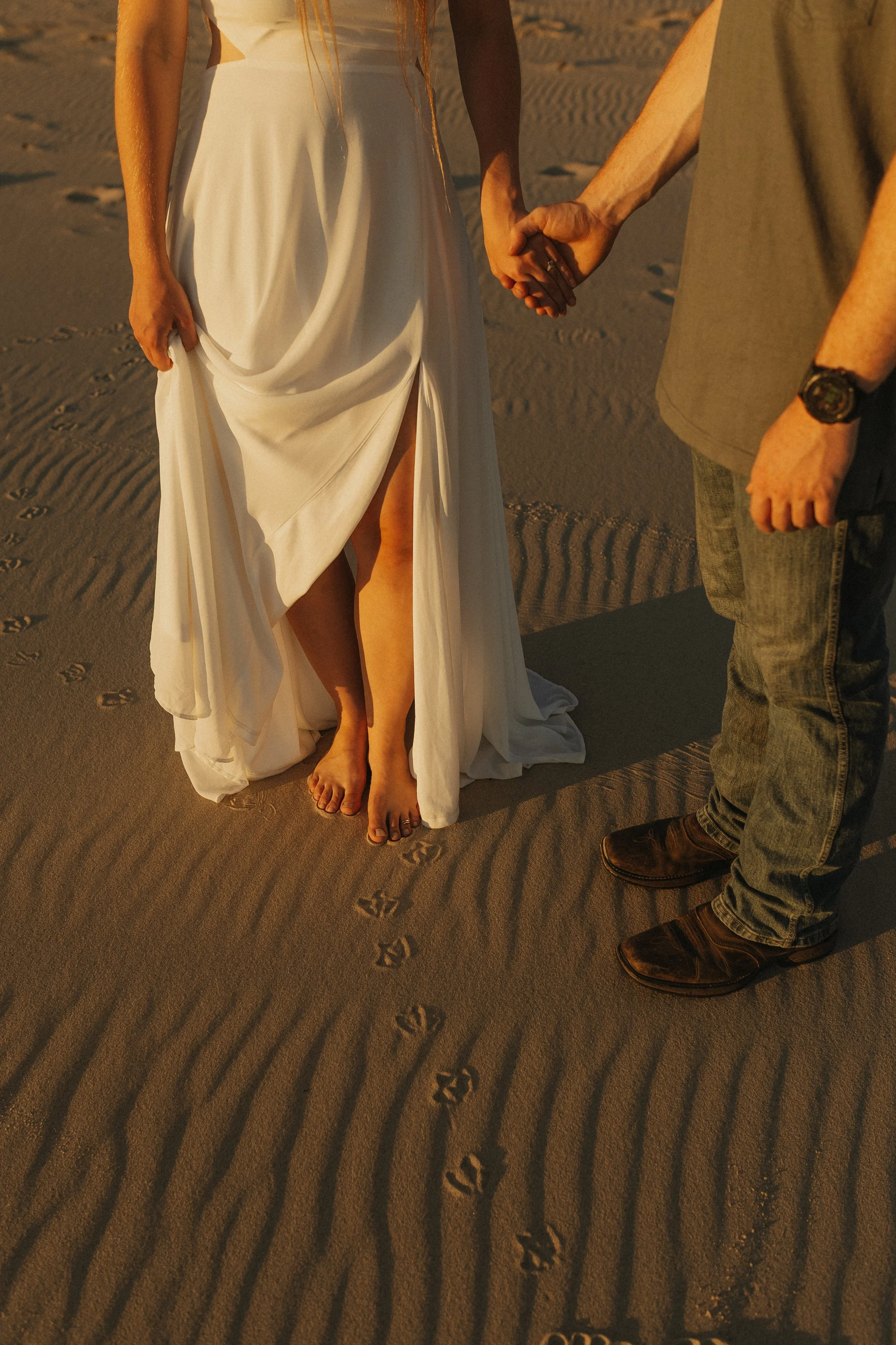 A couple holding hands standing on sandy beach with footprints in the sand.
