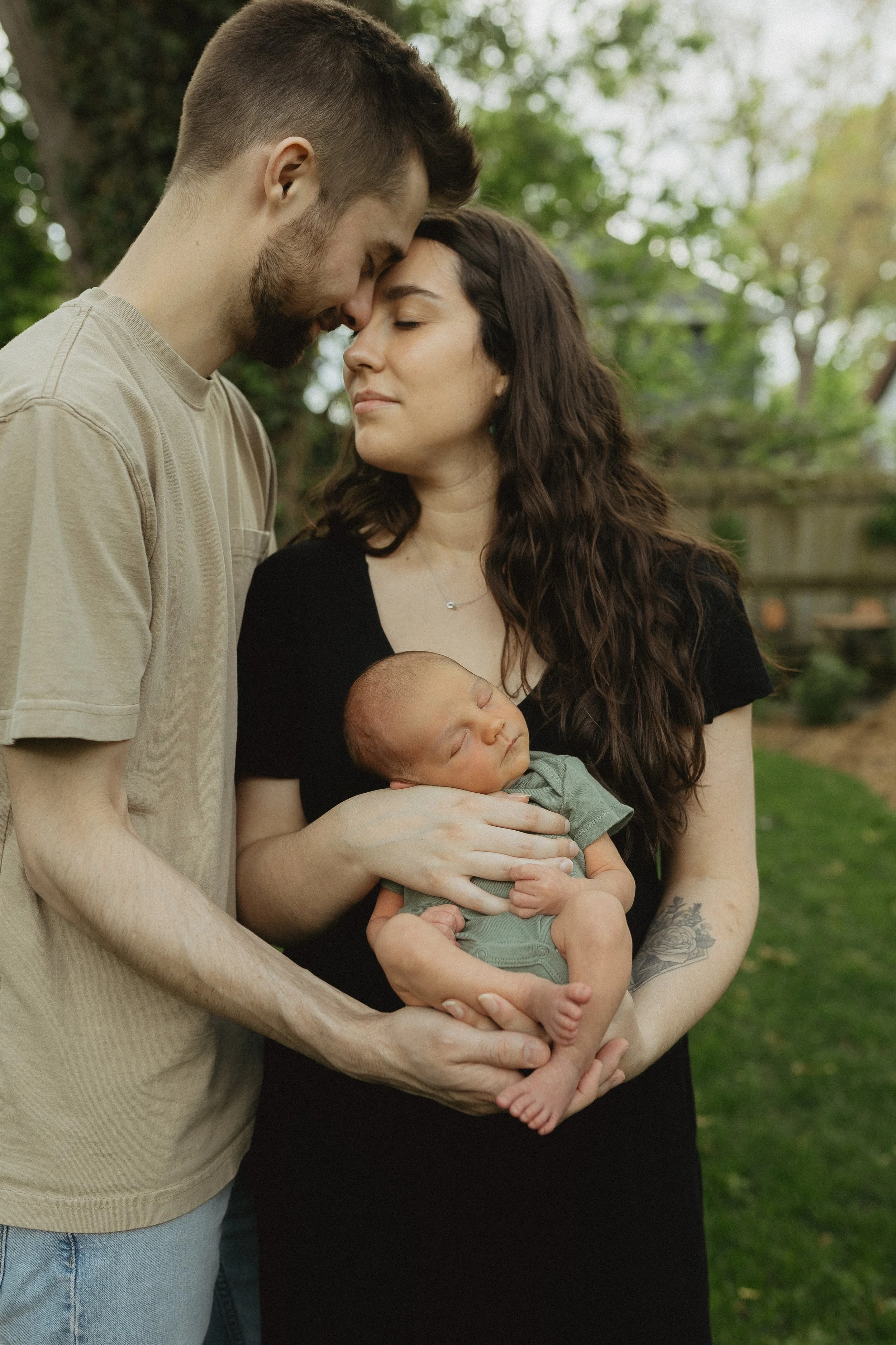 A family with a man, woman, and newborn baby outdoors, sharing an intimate moment with their foreheads touching.