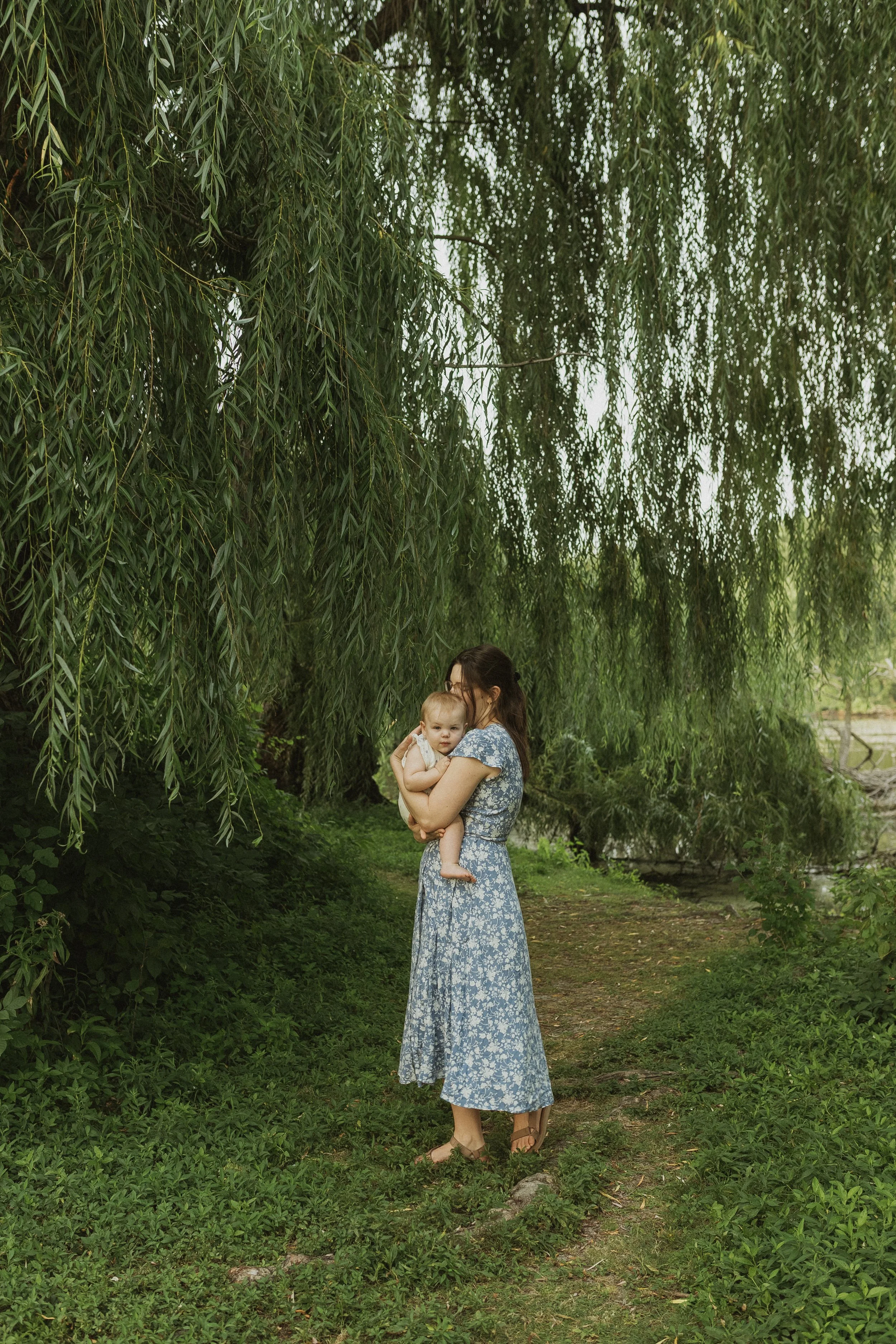 A woman in a blue floral dress holding a toddler in a green outdoor park with hanging tree branches.