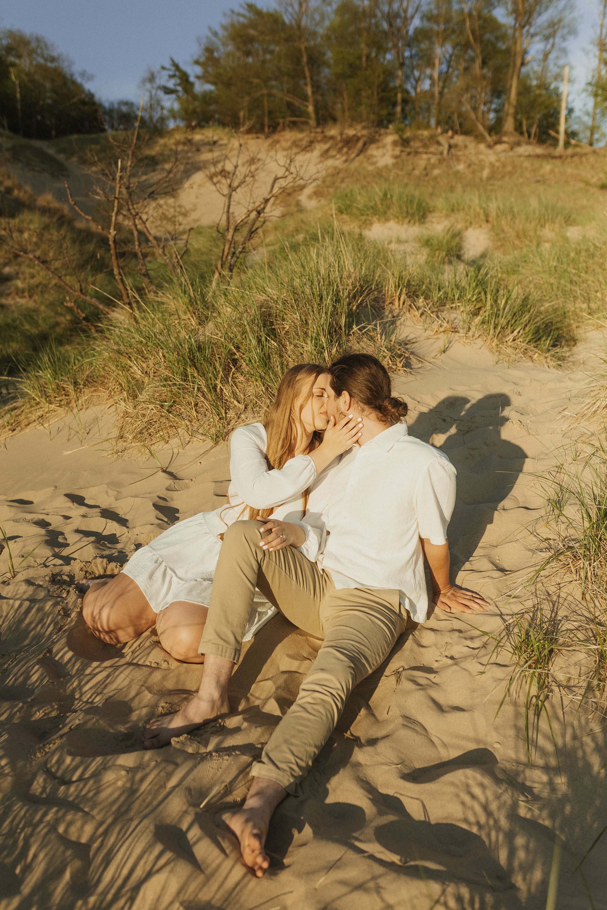 A couple sitting on a sandy beach, kissing, surrounded by sand dunes and grass, with trees in the background, during sunset.