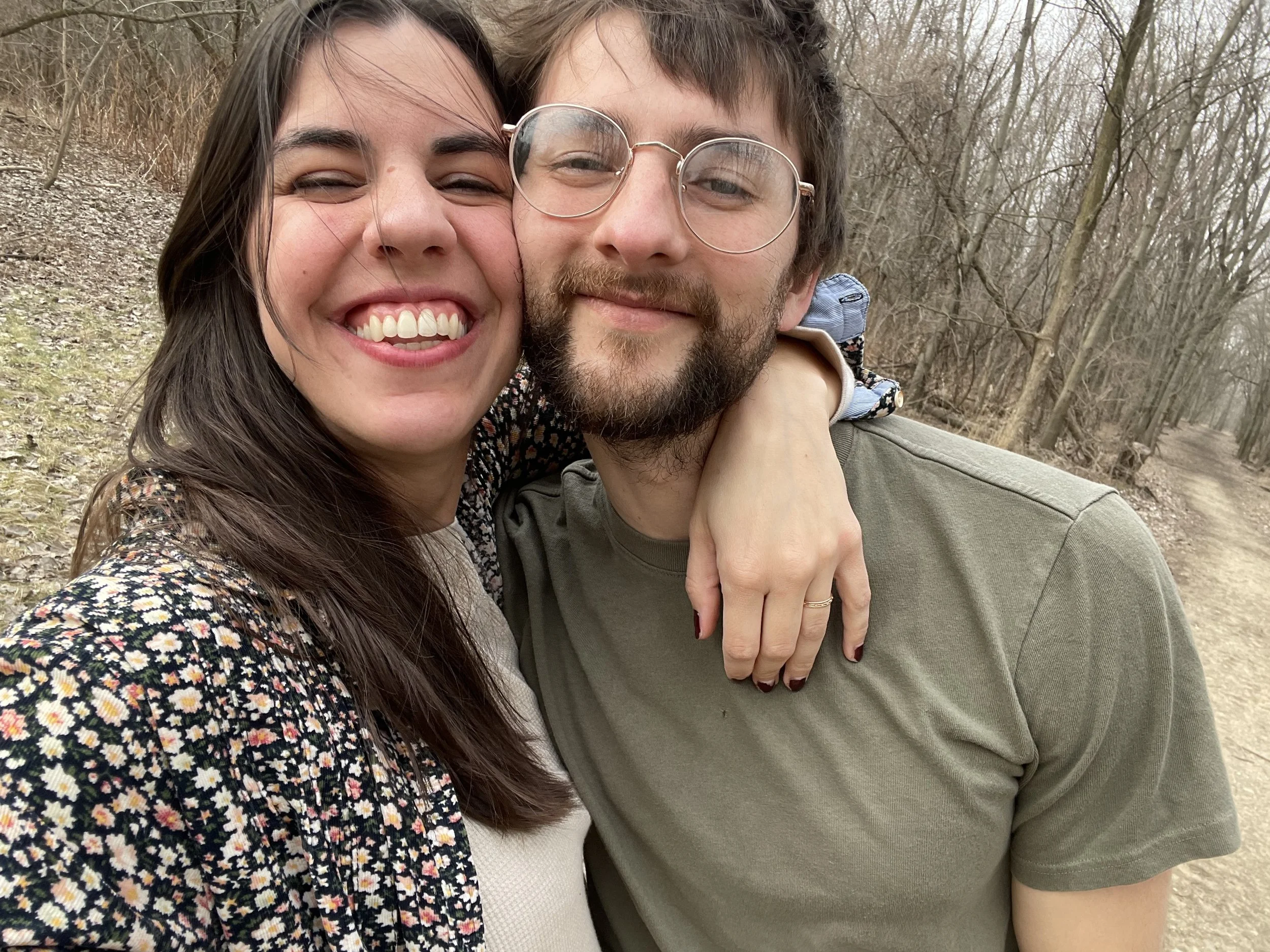 A smiling couple taking a selfie outdoors on a forest trail in early spring, with leafless trees in the background.