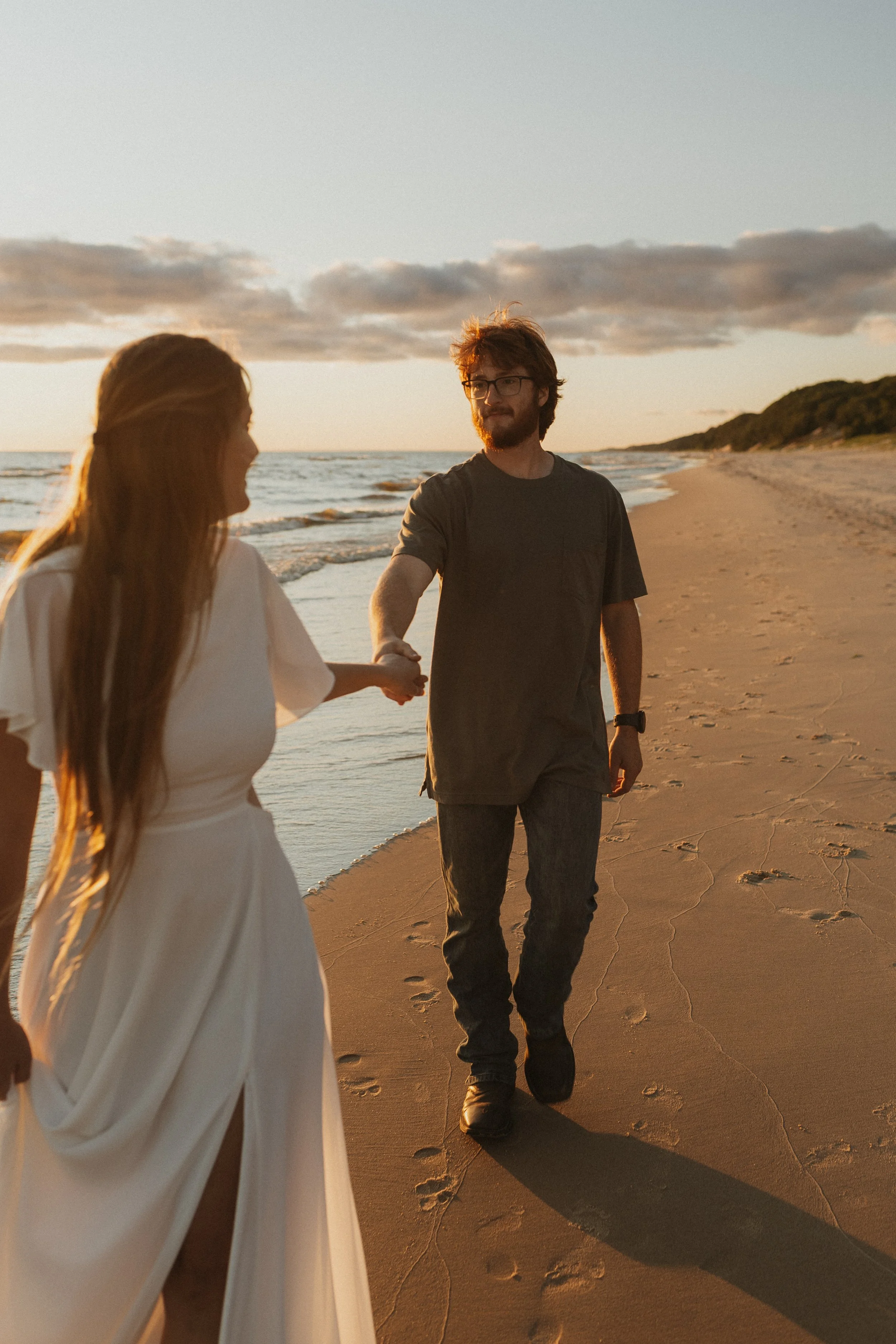 A man in a dark t-shirt and jeans walking on the beach, holding hands with a woman in a white dress during sunset.