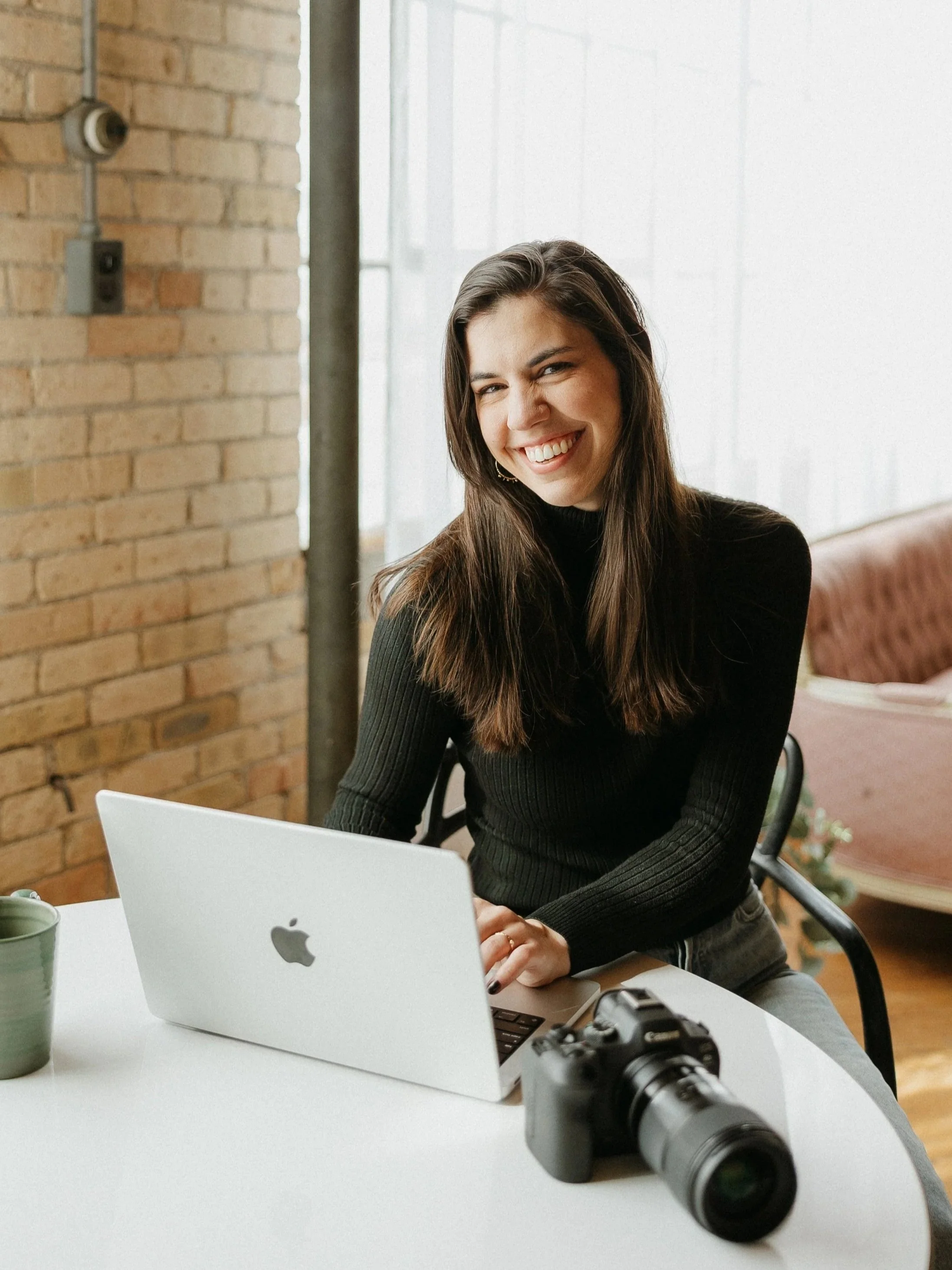 A woman with long brown hair, wearing a black sweater, sitting at a white table with a silver MacBook and a camera, smiling and winking at the camera in a room with a brick wall and large window.