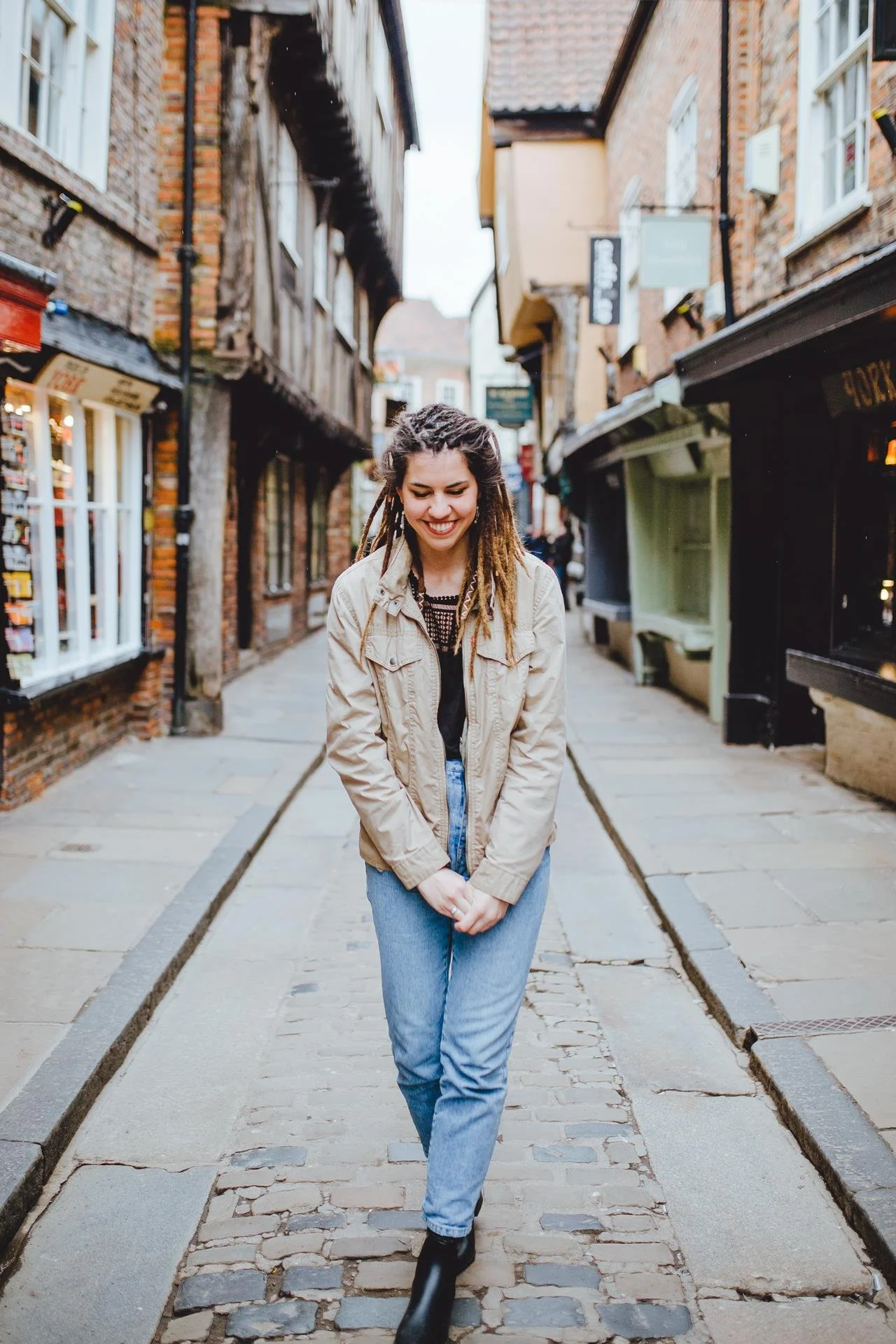 A young woman with dreadlocks walking down a cobblestone street in a historic area, smiling and looking down.
