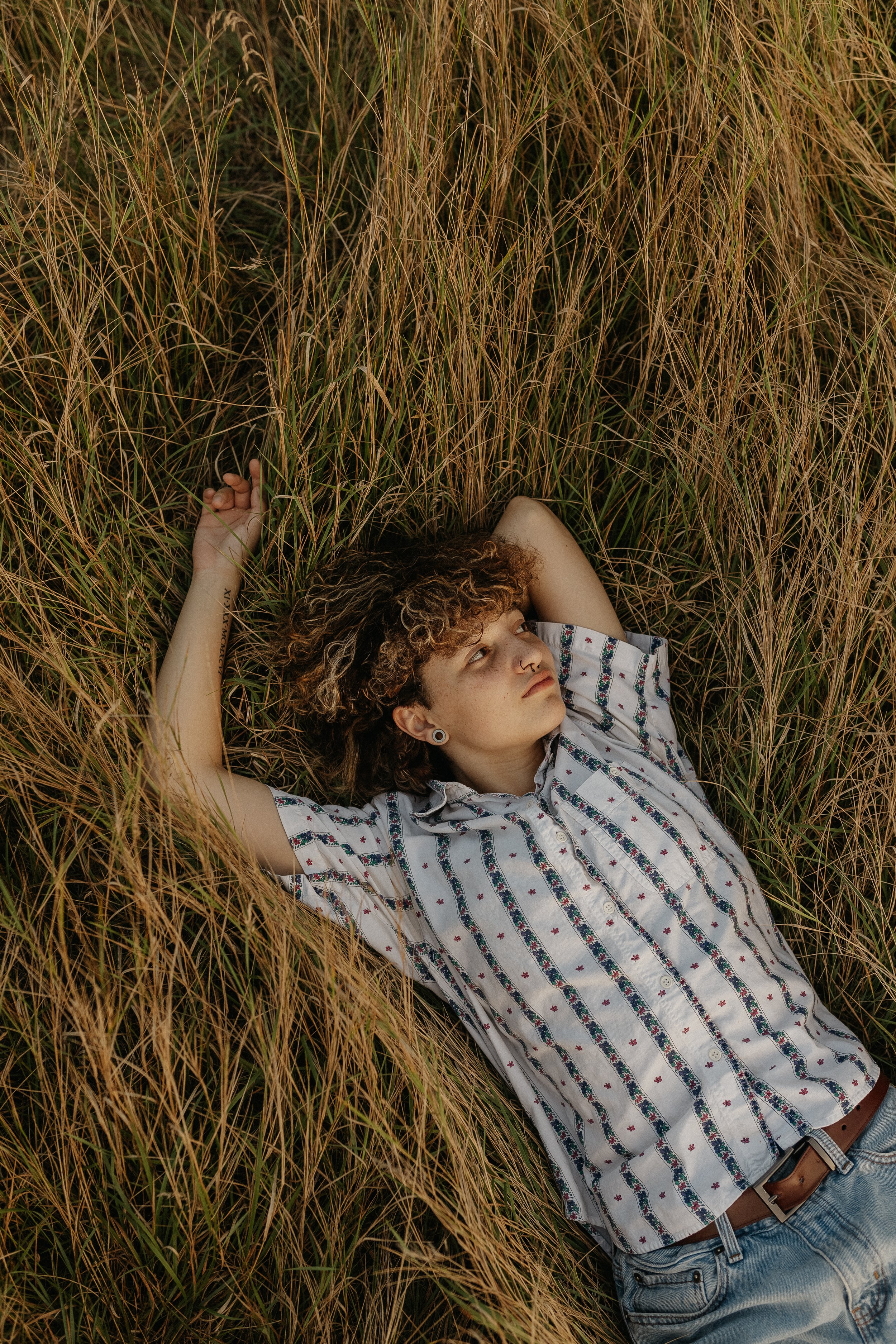 Person lying in tall grass, wearing a striped shirt and jeans, looking upward.