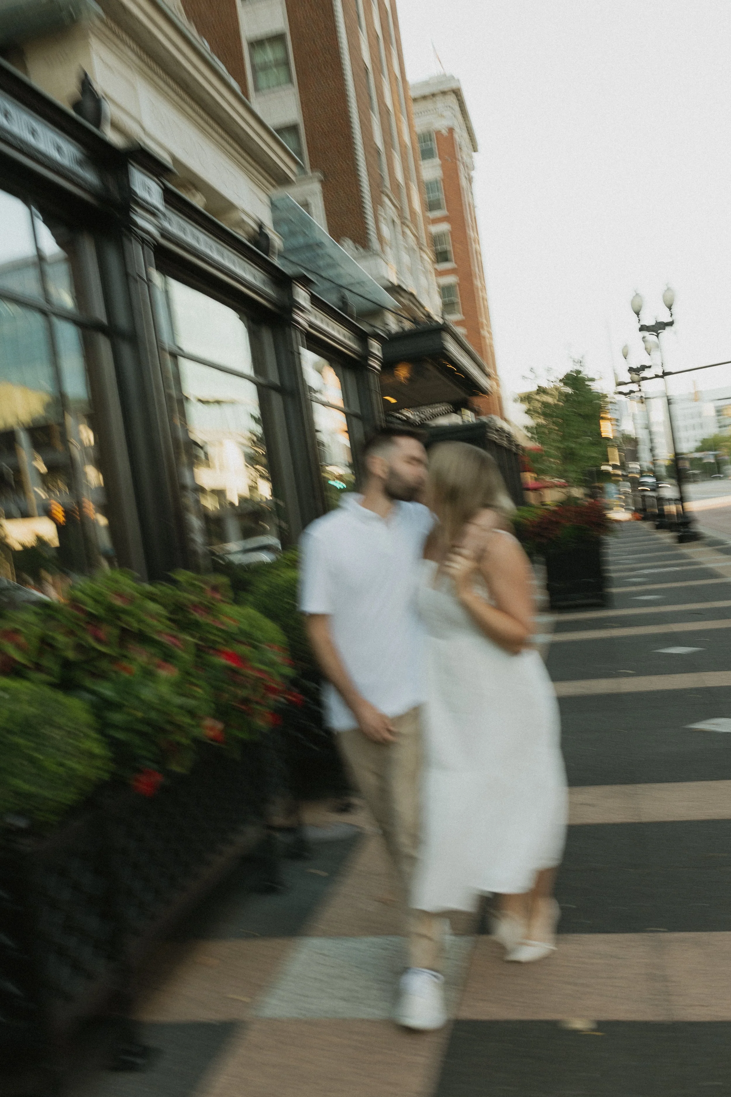 A blurry photo of a couple walking and kissing on a city sidewalk during the daytime, with buildings and street lamps in the background.