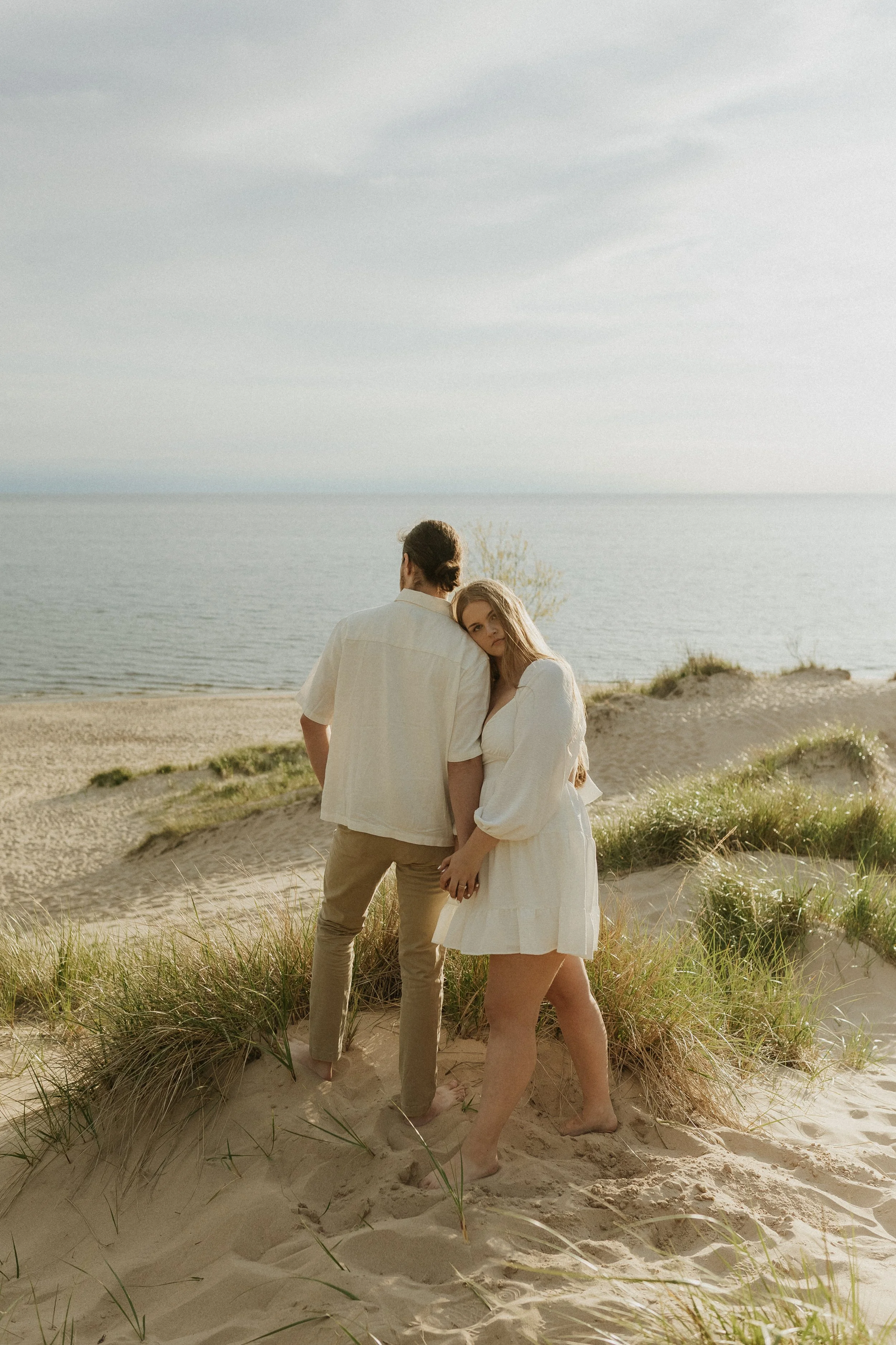 A young couple standing on a sandy beach with dunes and grass, overlooking the ocean during sunset. The woman is leaning her head on the man's shoulder, both dressed in light-colored clothing, creating a romantic and peaceful scene.