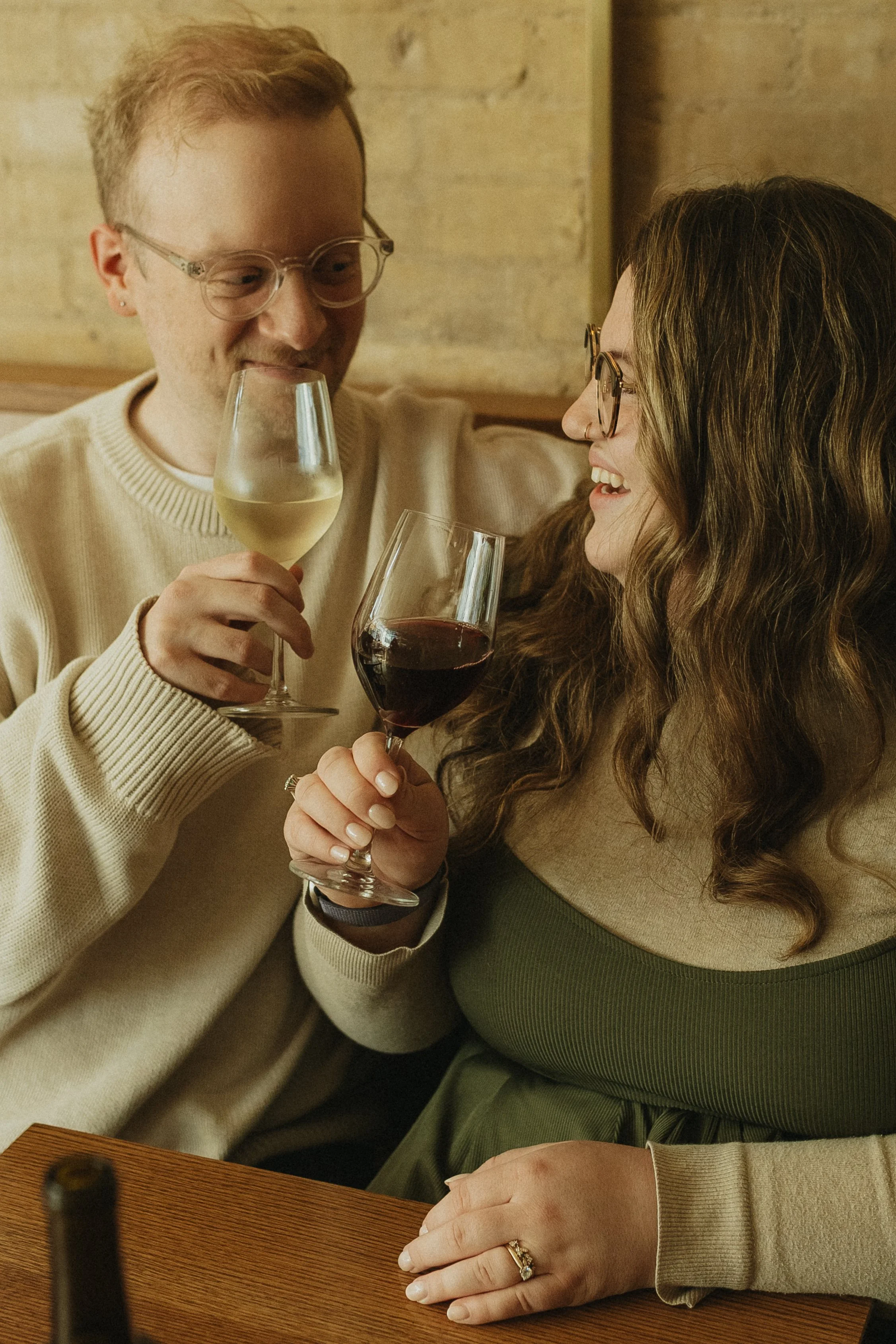 A man and woman sitting together, holding glasses of wine, smiling and enjoying each other's company in a cozy indoor setting.