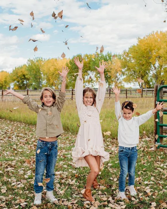 White Barn Acres | Northern Colorado Farm Photo Venue | three smiling young children throwing fallen leaves into the air in front of grassy field, split rail fence line and a row of golden cottonwood trees