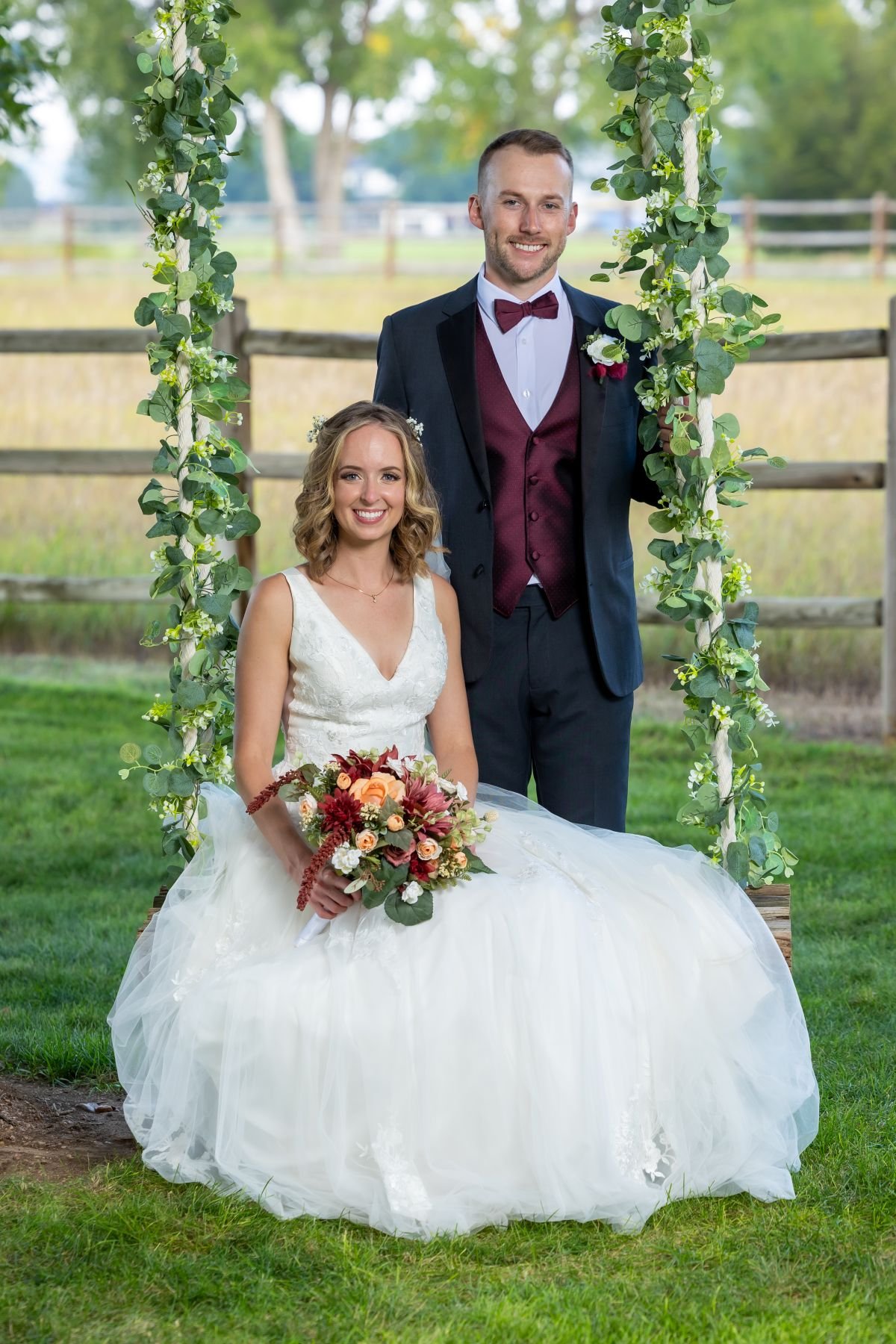 White Barn Acres | Northern Colorado Farm Photo Venue | smiling bride is sitting on a swing holding bouquet with smiling groom standing behind holding onto eucalyptus adorned swing ropes with green grass under feet and wooden split rail fence behind