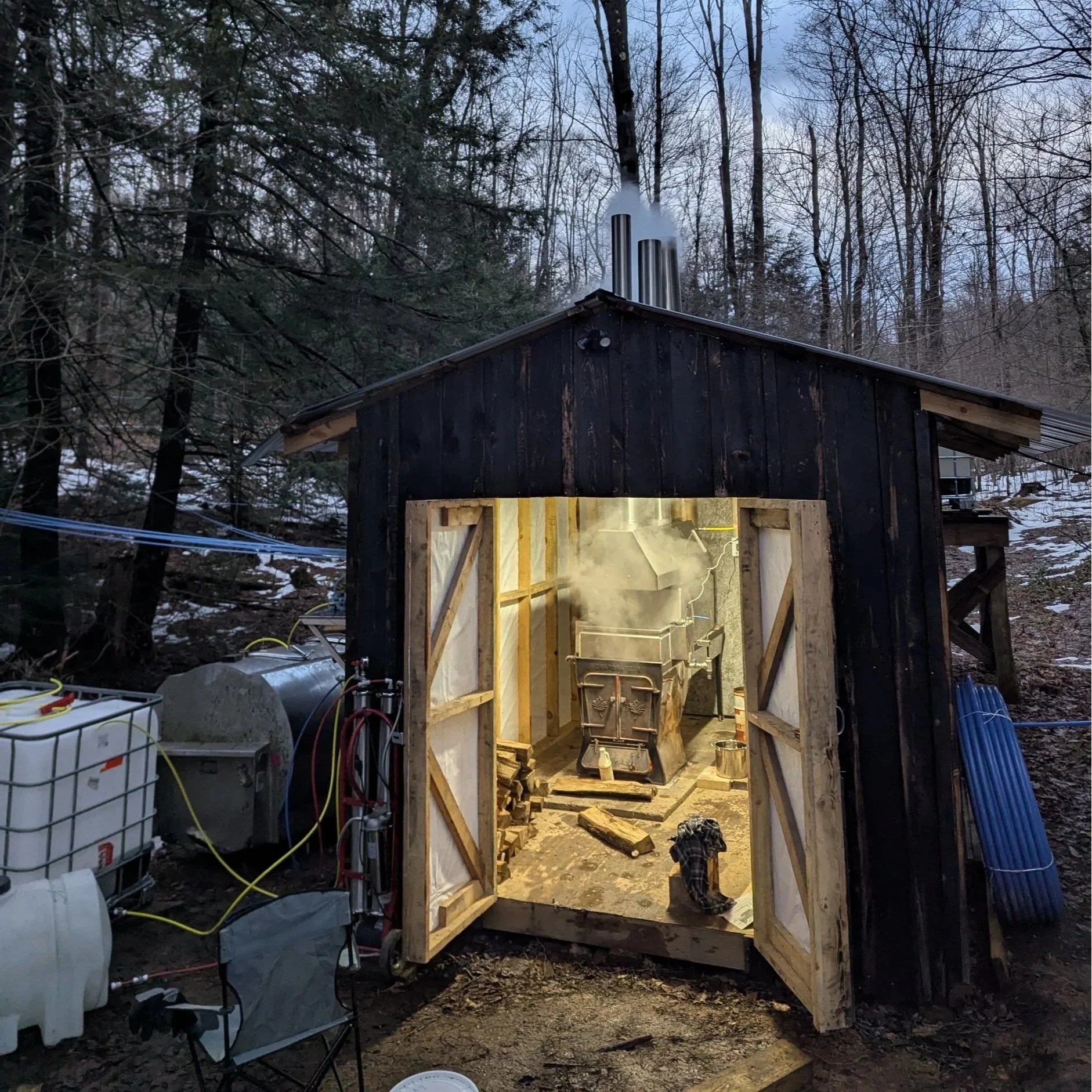 A black maple sugar shack in the woods with evaporator boiling maple syrup. Steam is coming out of the top exhaust stacks. Outside is maple sap storage tanks and equipment.