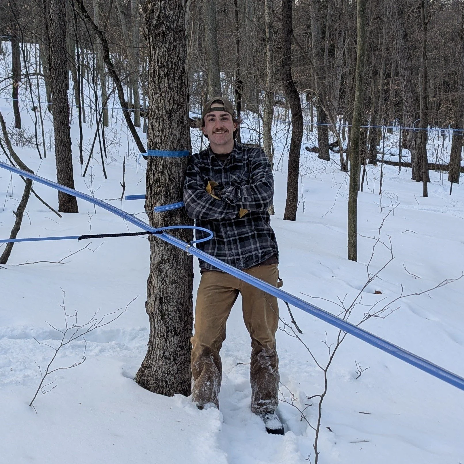 A man with a plaid shirt, tan pants, and a backwards cap standing in a snowy forest, smiling, with his arms crossed, leaning against a tree.