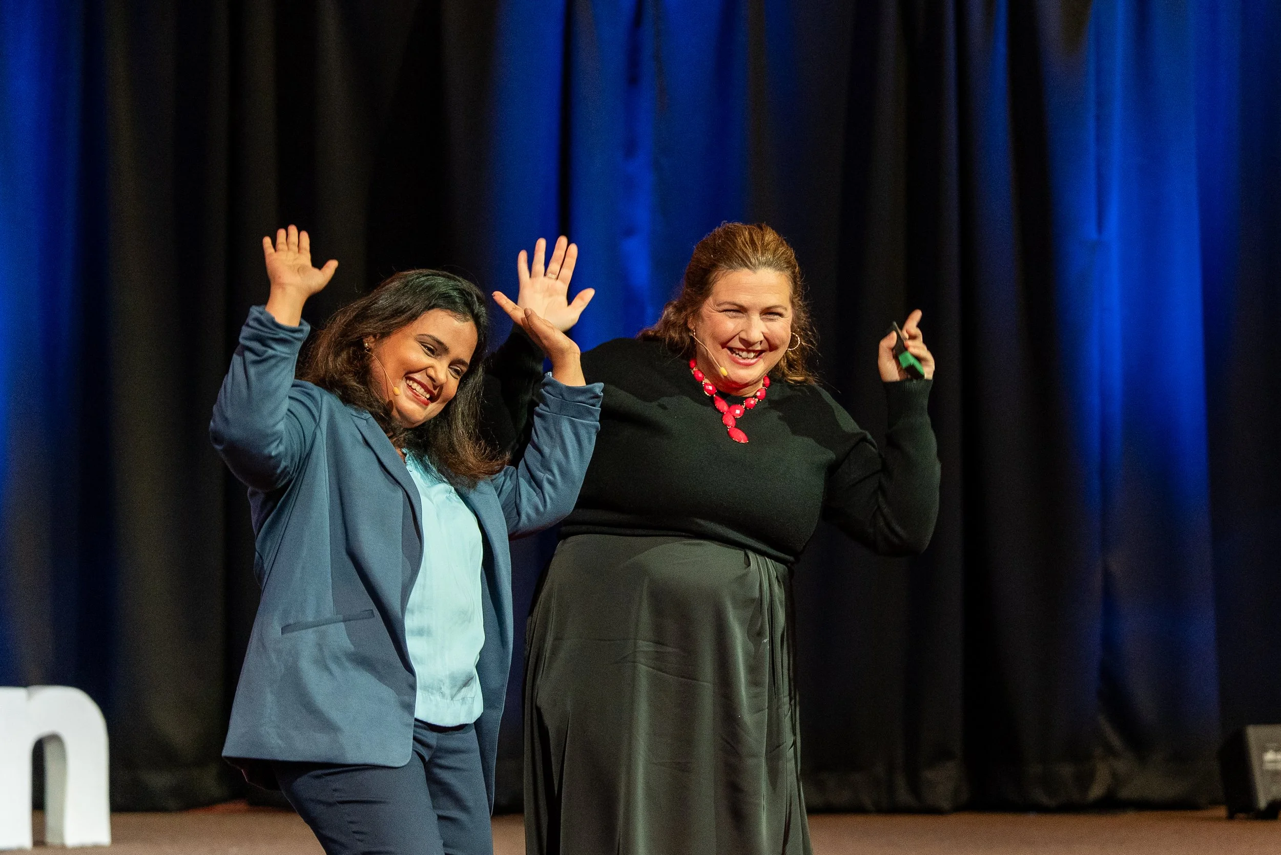 Two women standing on stage, smiling and waving with black curtains behind them.
