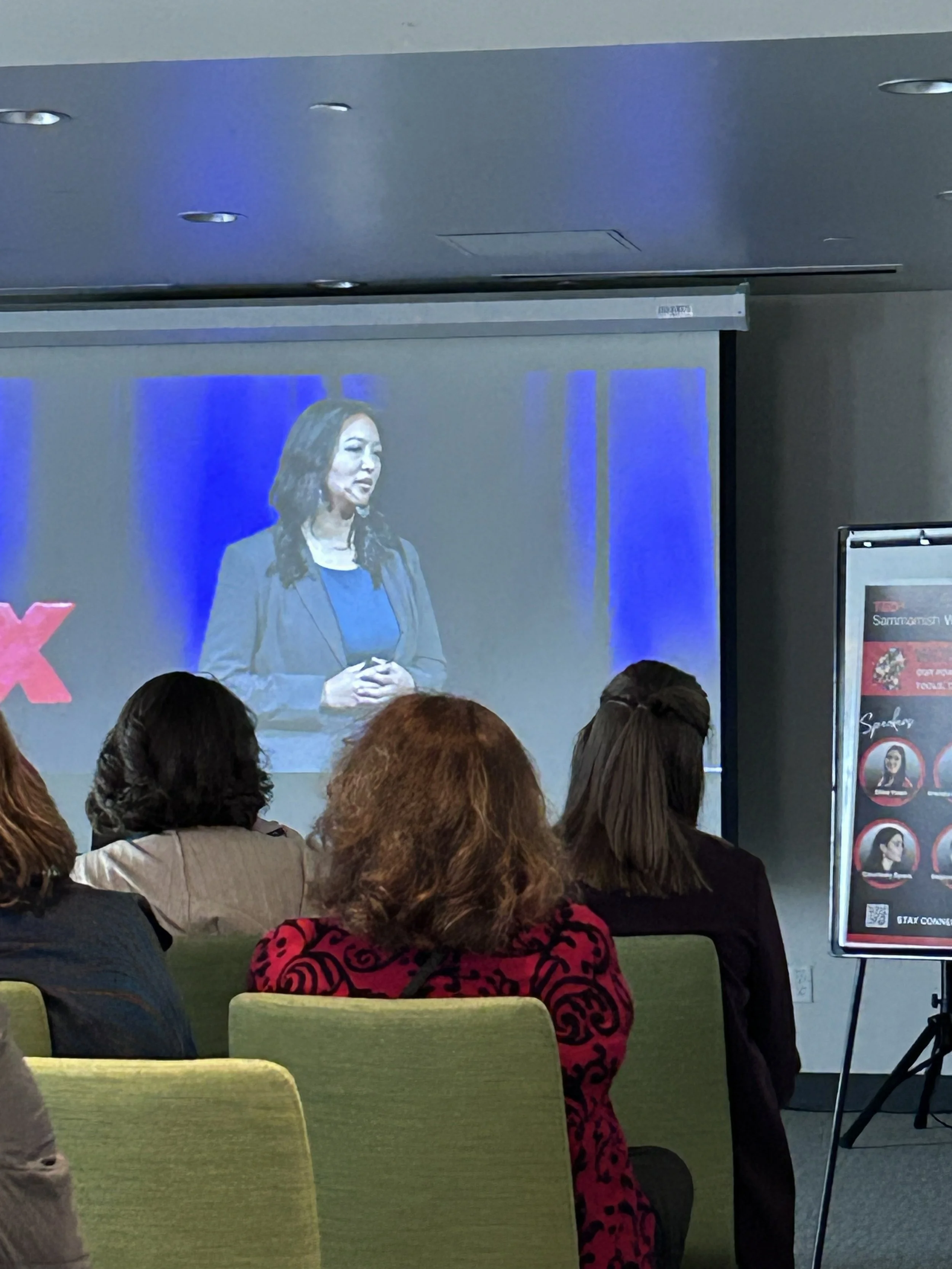 A woman in a blue blouse and gray blazer speaking on a stage during a presentation or conference, with an audience seated and watching her.