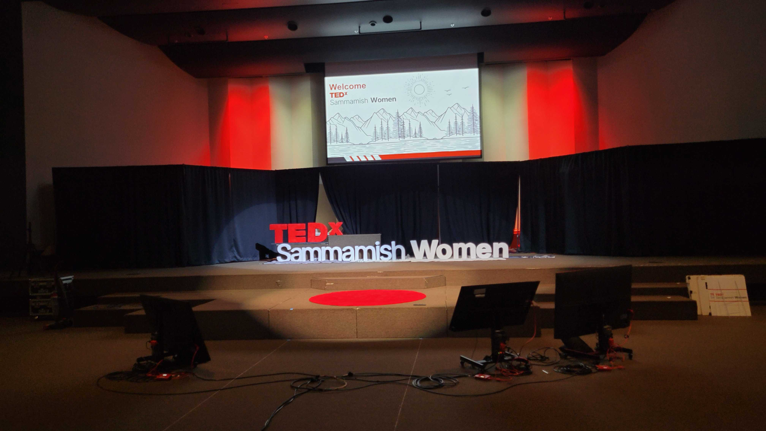 Empty stage set up for a TEDx event with a large screen displaying a background with mountains, trees, and a sun, and text welcoming attendees to TEDx Sammamish Women. The stage features a TEDx sign and a sign displaying the event name and theme.