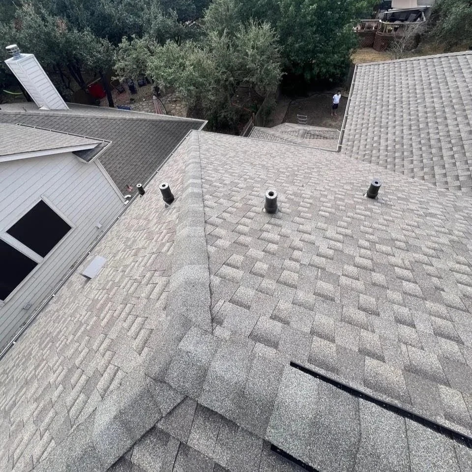 View of a residential rooftop with multiple sections covered in gray shingles, three vent pipes, and a white window on the side of a house. In the background, trees and a backyard with a person standing near a ladder are visible.