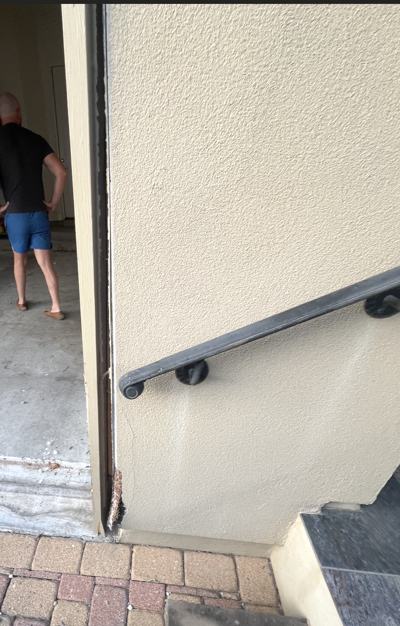 An exterior staircase with a black handrail attached to a beige wall, leading upward. Part of the building's entrance and a person in black shirt and blue shorts are visible inside the building.