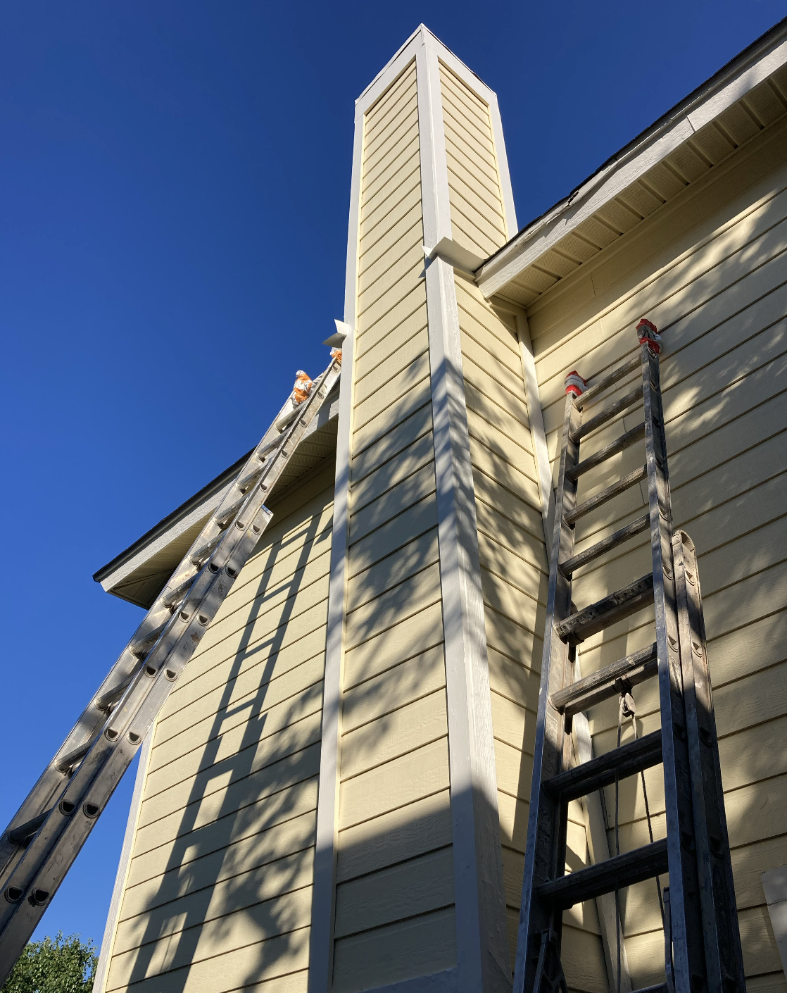 The exterior of a house under renovation showing two ladders leaning against a beige siding wall, with a chimney in the center, against a bright blue sky.