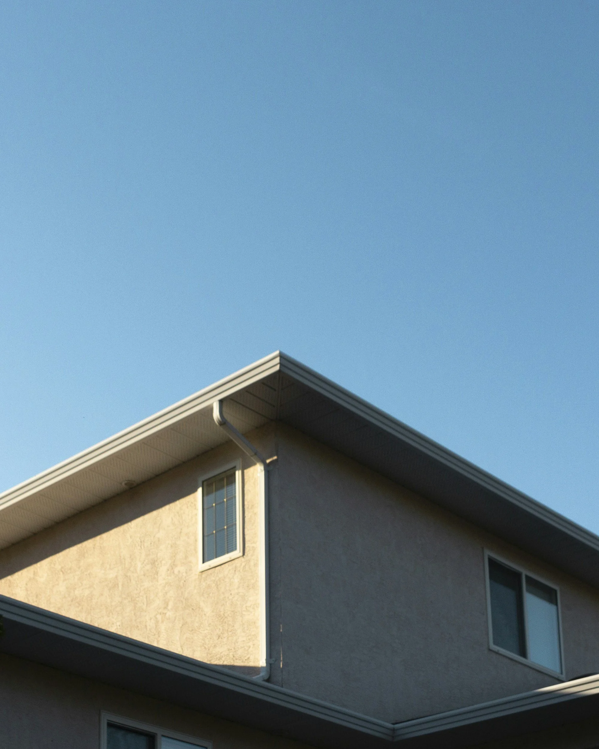 The corner of a multi-story residential house with beige stucco exterior walls, white trim, and a sloped roof, set against a clear blue sky.