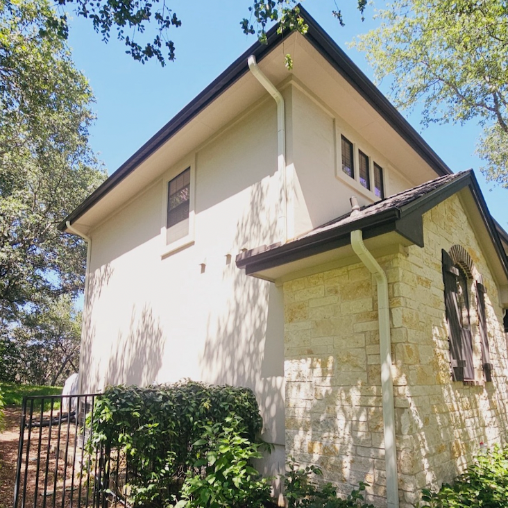 Side view of a two-story house with a cream-colored stucco exterior, stone accents, black shutters, and a sloped roof with gutters, surrounded by trees and greenery.