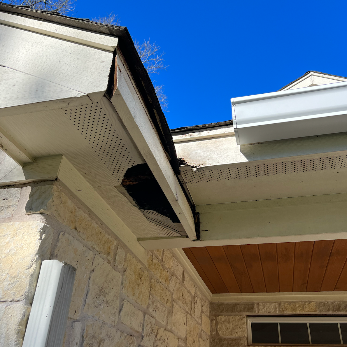 Close-up of a damaged section of house eaves showing rotted wood, with visible deterioration and peeling paint. The house has a stone exterior, a gutter system, and a clear blue sky in the background.