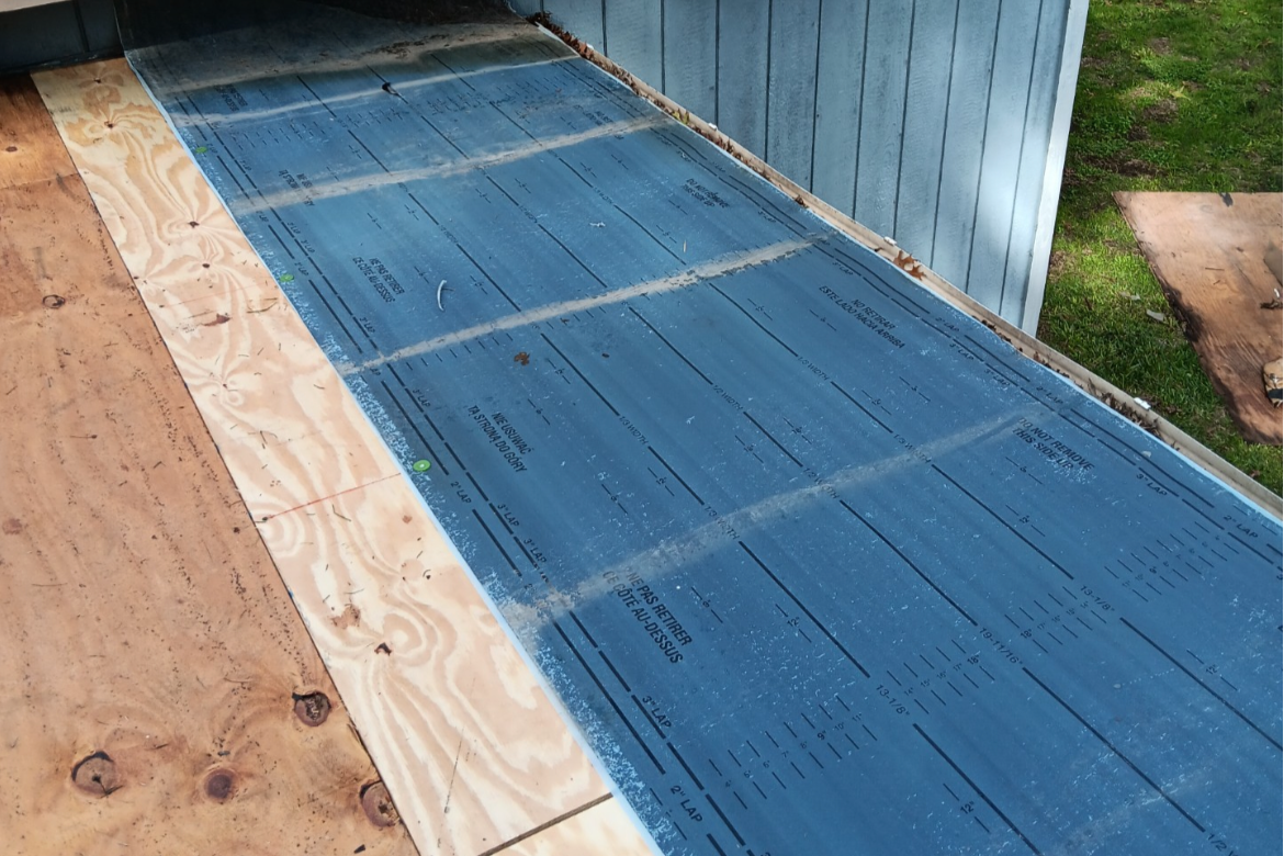 Construction site with plywood flooring and a blue vapor barrier or roofing underlayment installed on the subfloor, next to a blue exterior wall.