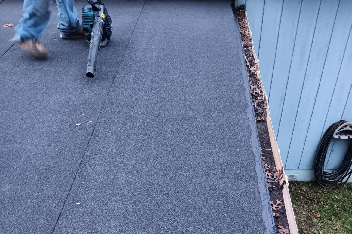 Close-up of work being done on a roof, showing a person operating a leaf blower and a section of the roof with a new waterproofing layer. The wall of a house with wood siding and some gardening tools are visible.