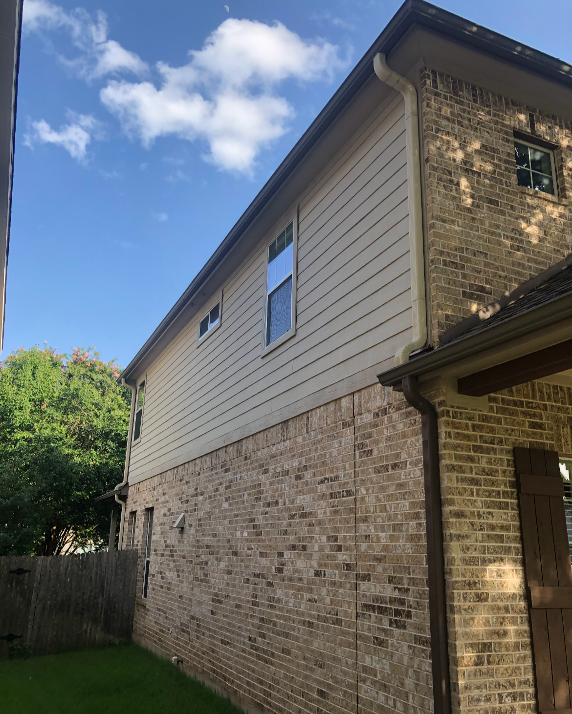 The side of a two-story house with a brick lower level and light-colored vinyl siding upper level, featuring three windows, gutter systems, and surrounded by greenery and a blue sky with a few clouds.