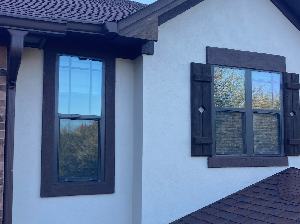 Close-up of a house's exterior showing two windows with dark wooden trim and shutters, beige stucco walls, and a brown shingled roof.