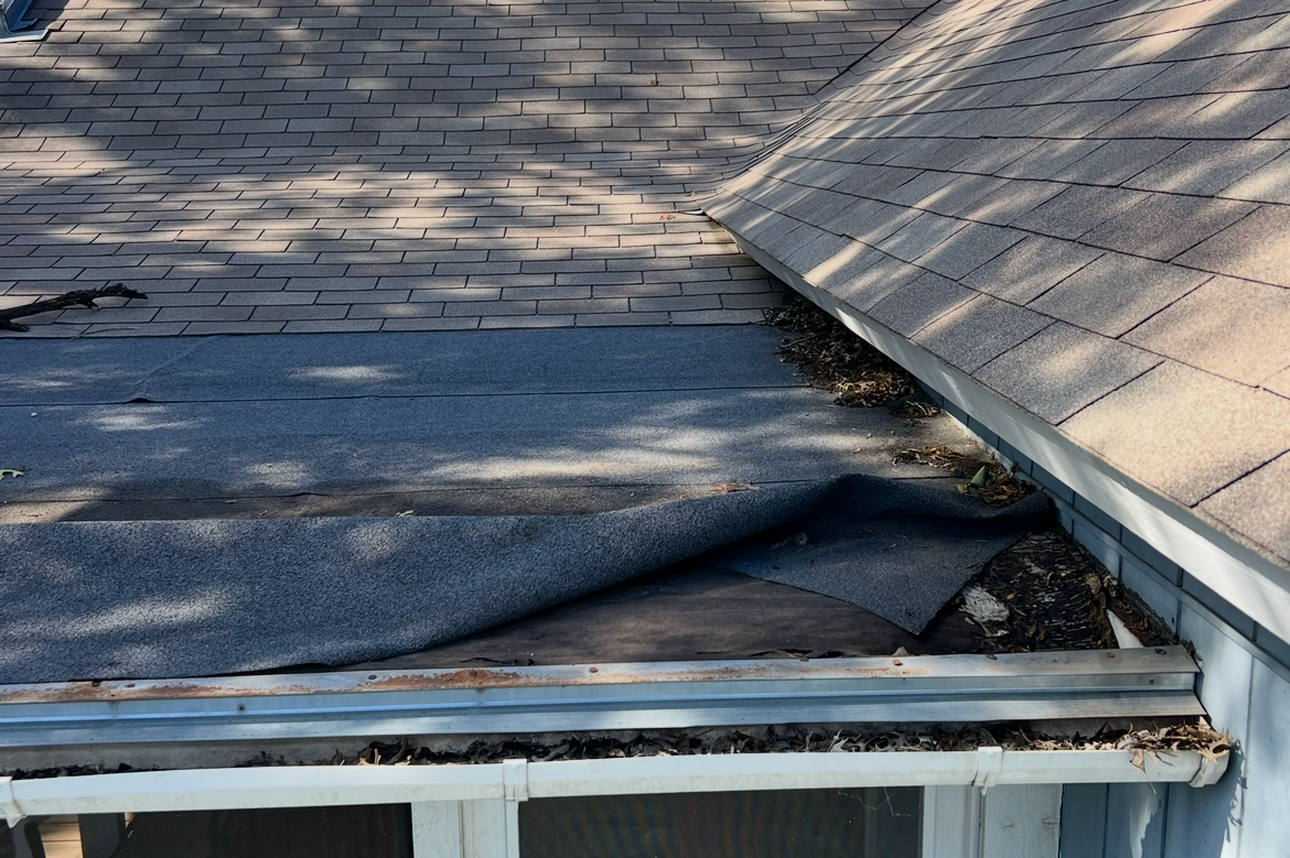 Close-up of a roof with worn shingles and a patch of weathered, rolled roofing material showing signs of damage and debris.